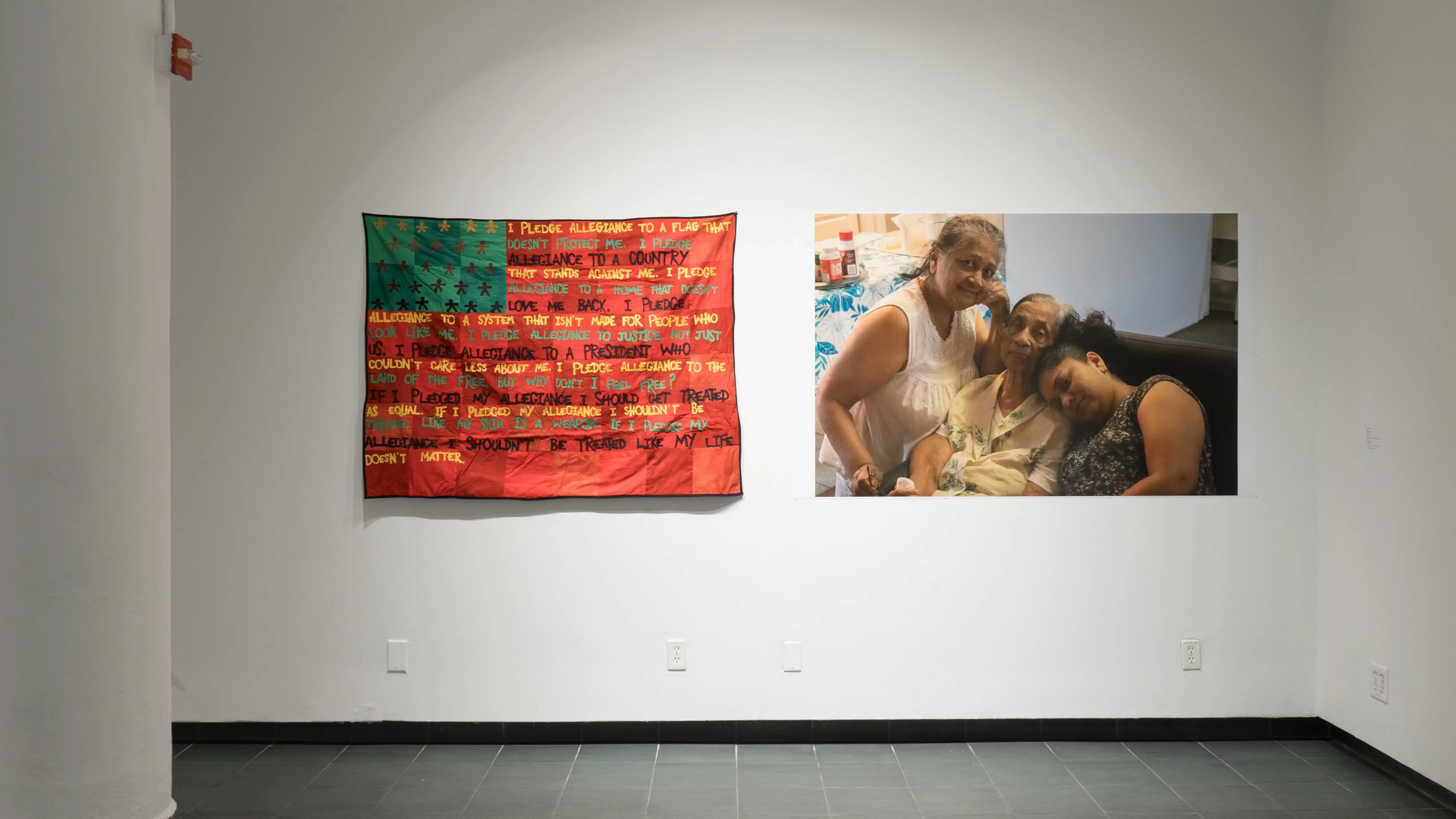  A closer view of one wall in the back gallery, showing a large textile work resembling a flag with words as stripes, and a large print of three figures embracing and looking at the camera.