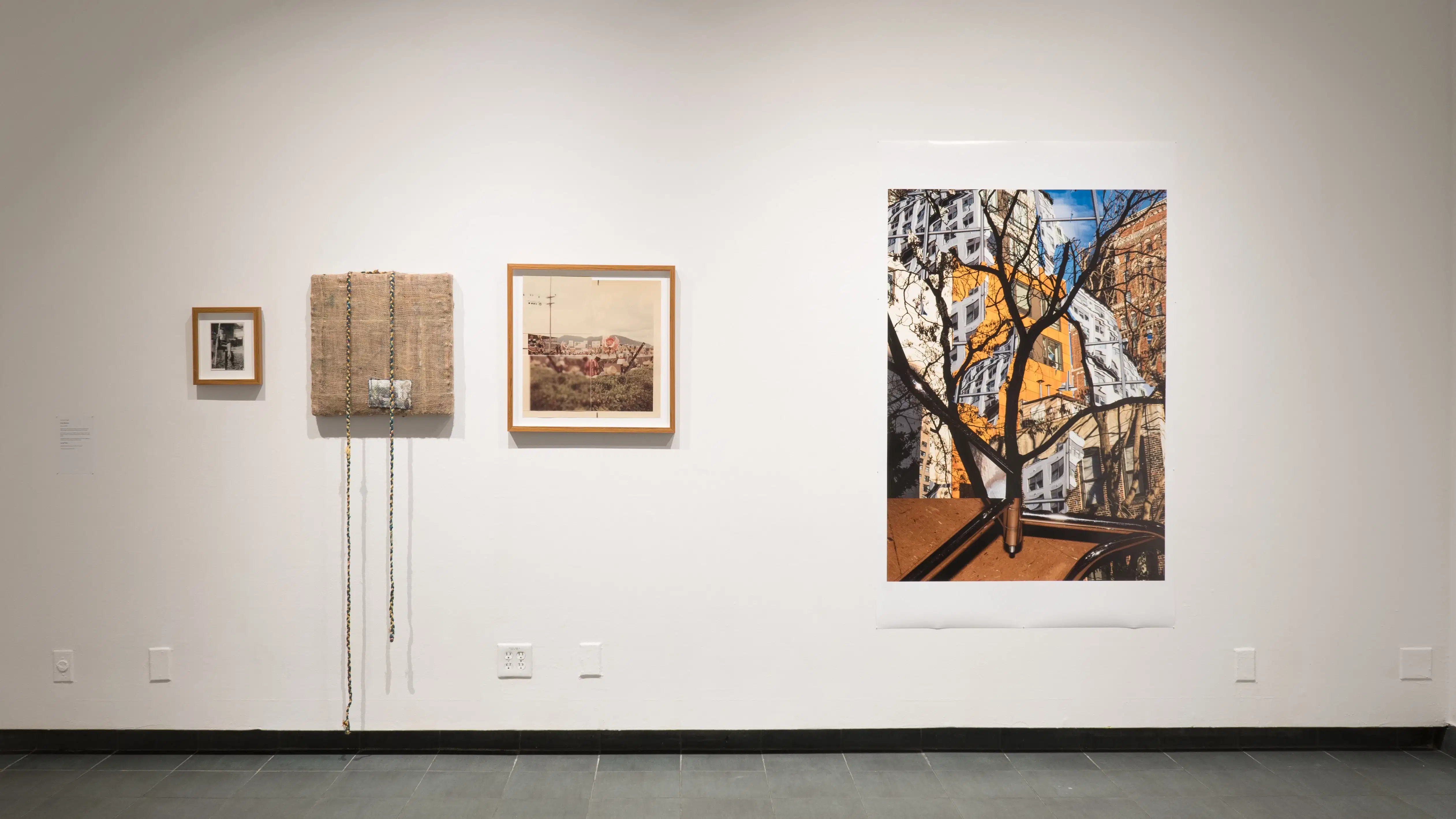 A closer view of one wall in the back gallery, showing three works on the left—two framed and one textile work—and a large print of a cityscape on the right.