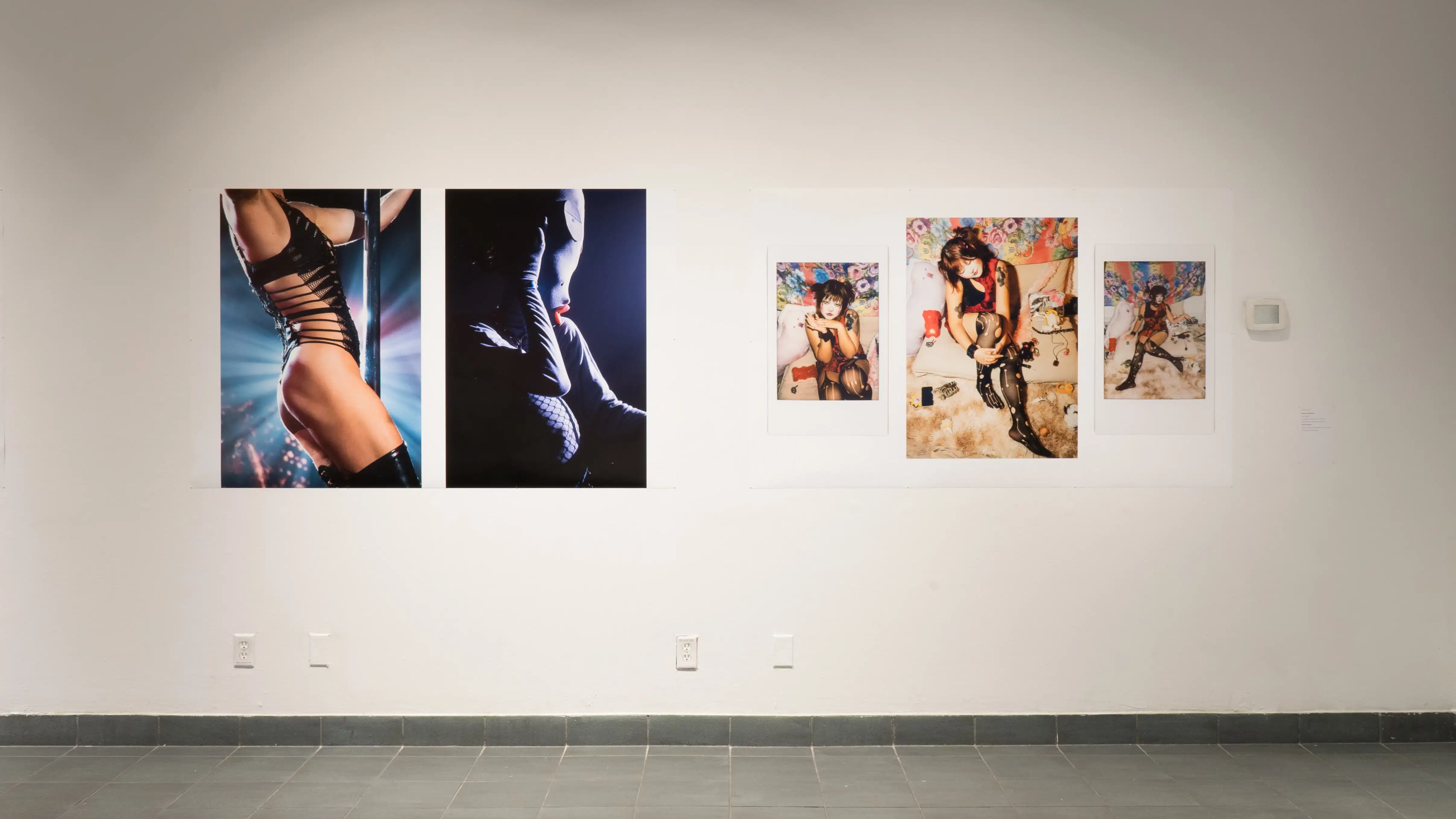 A closer view of one wall in the back gallery, showing two large prints: the left has two human figures, one in lingerie and one in a head-to-toe body suit in dancing poses; the right has three images of a feminine human figure in repose on a bed with a flower textile hanging behind them.