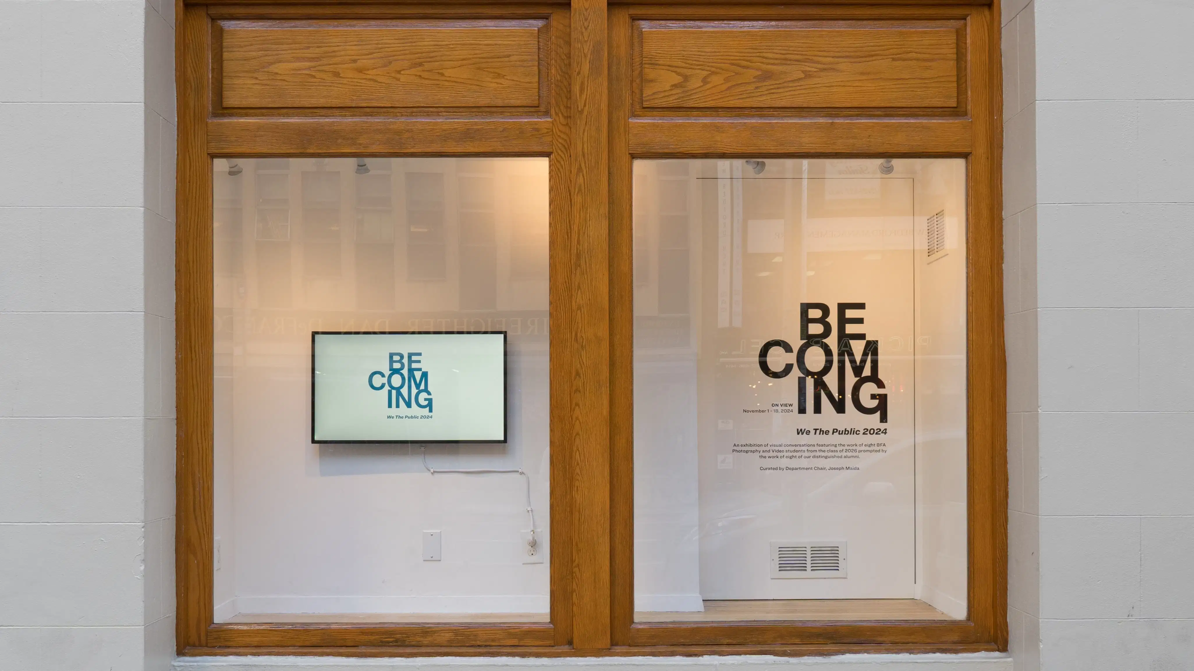 A view of the Gramercy Gallery window from the street, with a monitor showing the word “Becoming” on the left, and the same text on the right in vinyl adhered to the wall.