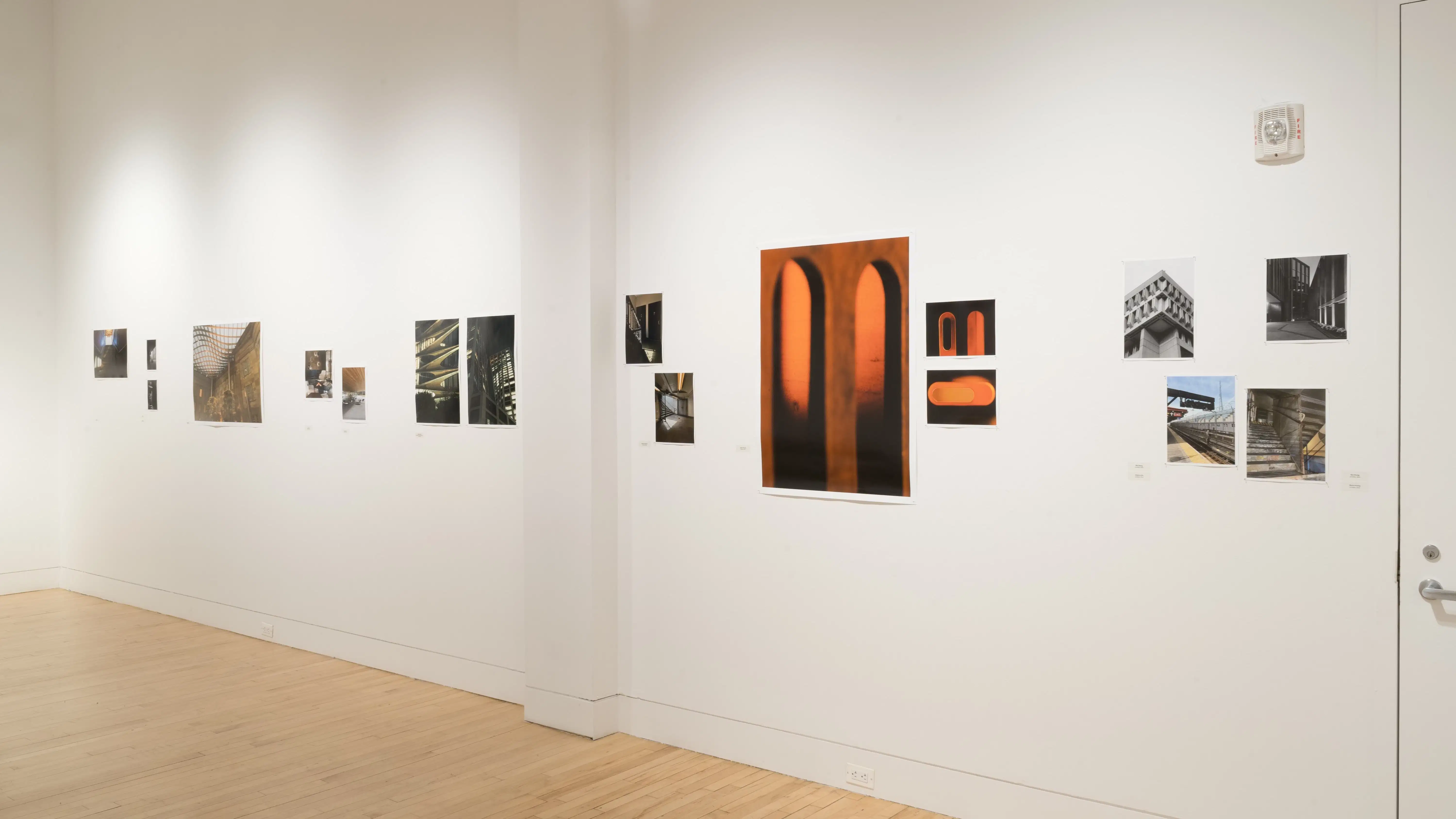 Angled shot of one wall in the Flatiron Gallery, showing multiple prints of industrial moments in varying sizes lining the walls, and an extra large print in orange showing two arches in the foreground..