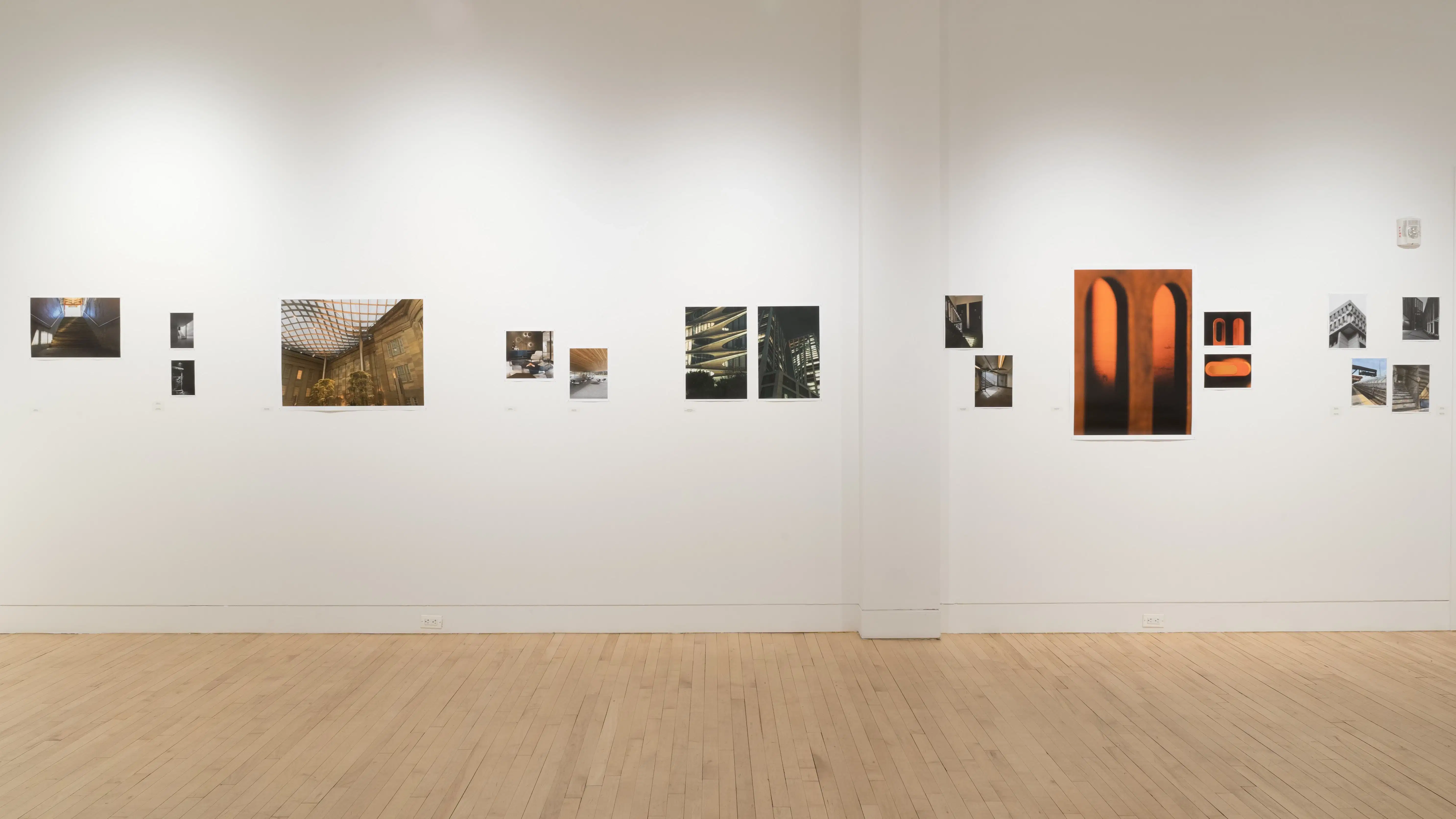 Straight shot of one wall in the Flatiron Gallery, showing multiple prints of industrial moments in varying sizes lining the walls, and an extra large print in orange showing two arches towards the right.