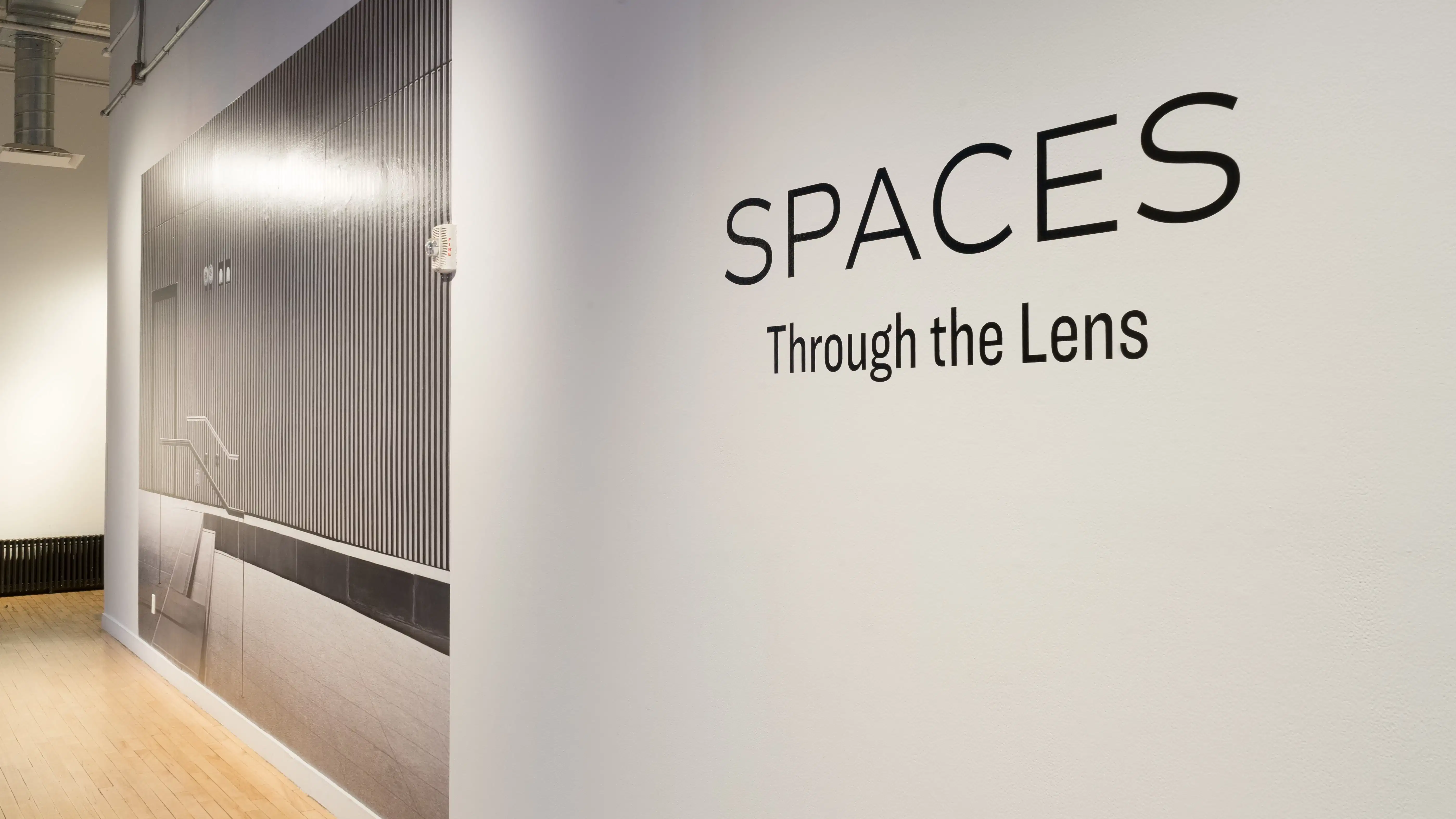 Angled shot of the window wall at the Flatiron Gallery, showing vinyl in large letters spelling "Spaces Through the Lens," and a large print covering the entire hallway wall of a grey industrial space.