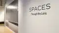 Angled shot of the window wall at the Flatiron Gallery, showing vinyl in large letters spelling "Spaces Through the Lens," and a large print covering the entire hallway wall of a grey industrial space.