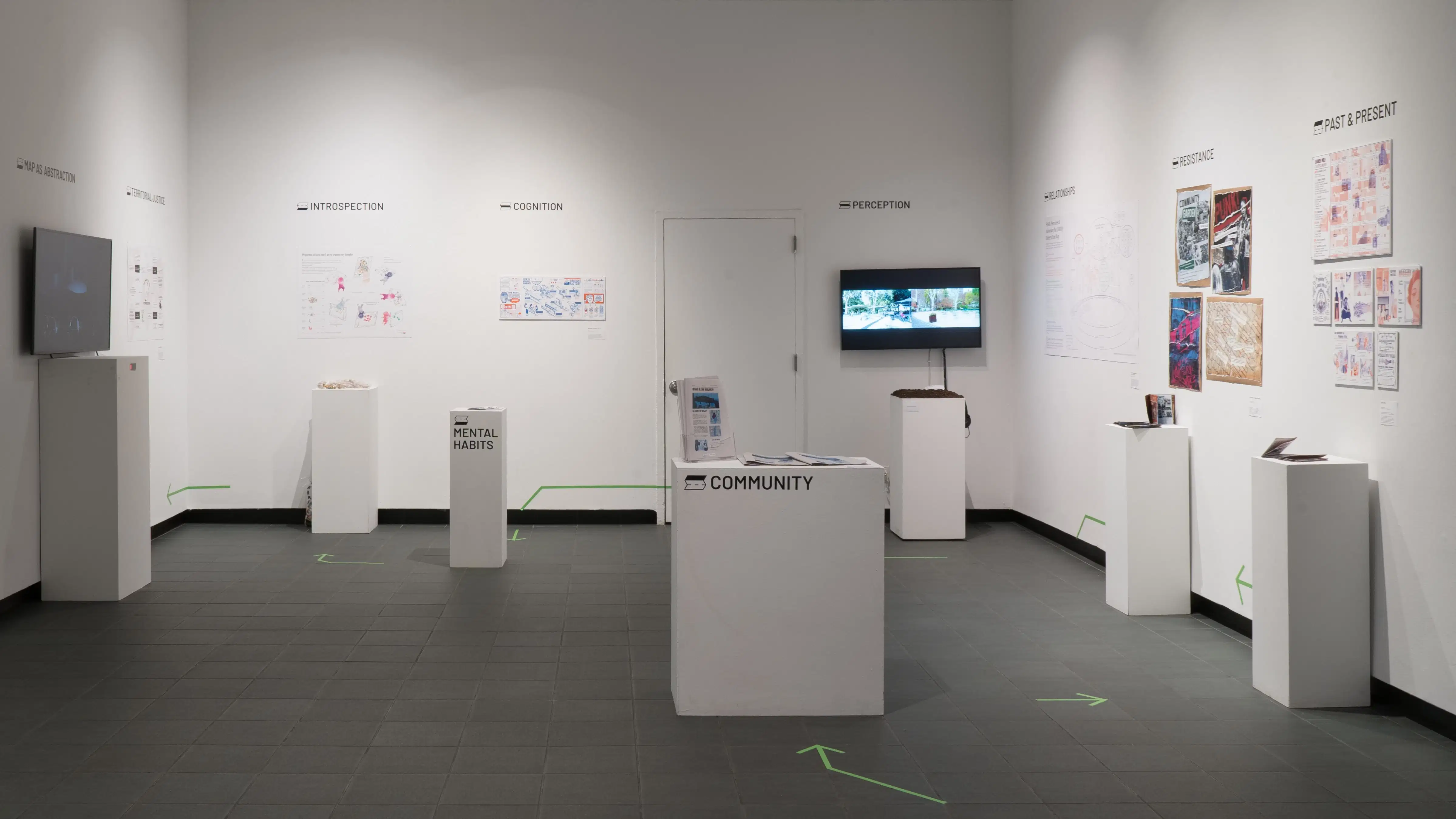 Green vinyl arrows lead visitors on a path through the gallery. Each stop at a pedestal or section of the wall has a title, such as “community” and “introspection.”