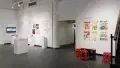 Graphics, posters, and monitors are displayed on the walls of the gallery space. Some objects sit on pedestals, and a few books lay on a small table surrounded by stools.