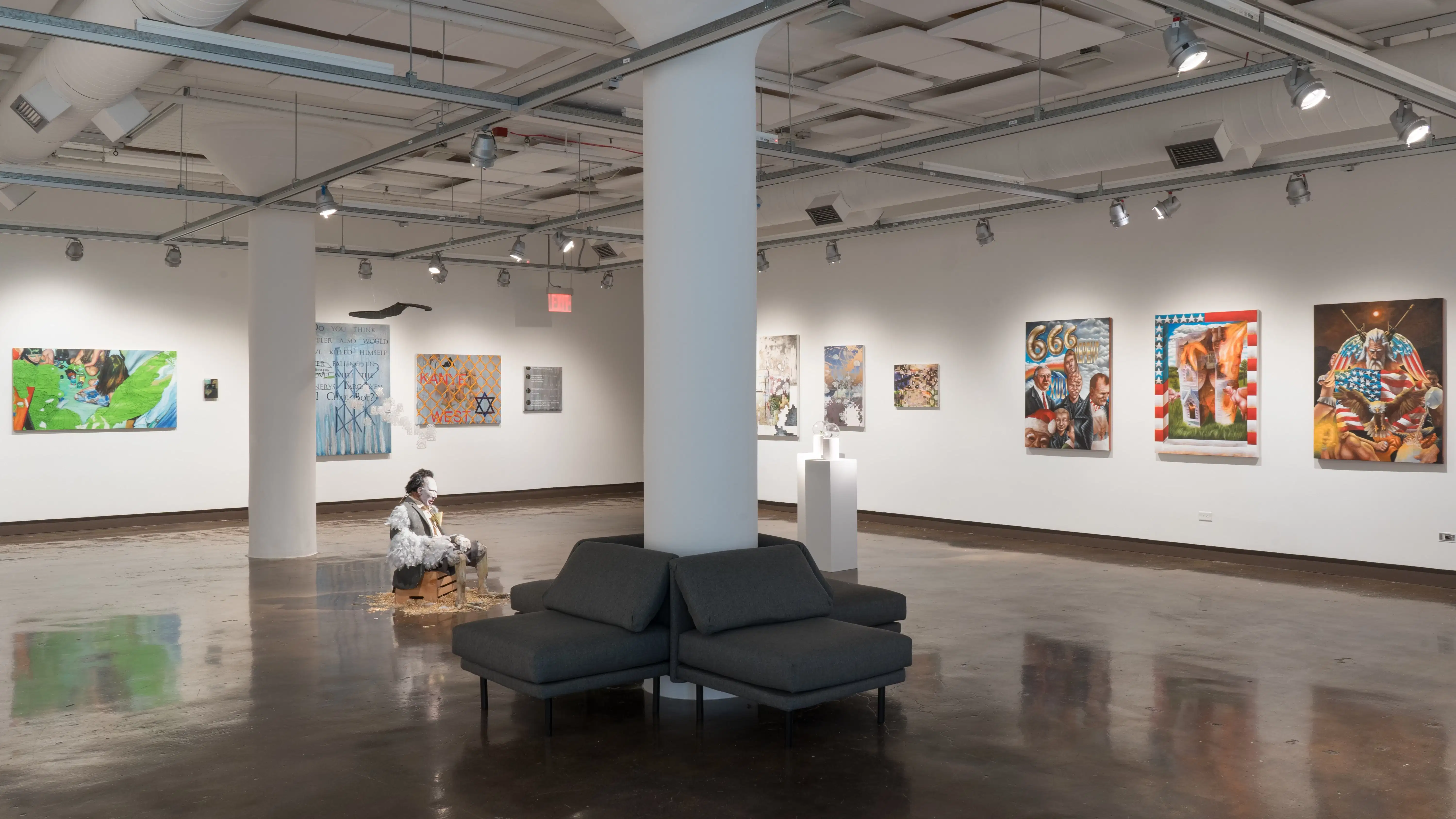 Large paintings and mixed media work on canvas line the walls of gallery one. A sculpture of a man with feathers, a hanging installation, and two pedestals are present in the middle of the gallery.