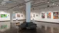 Large paintings and mixed media work on canvas line the walls of gallery one. A sculpture of a man with feathers, a hanging installation, and two pedestals are present in the middle of the gallery.
