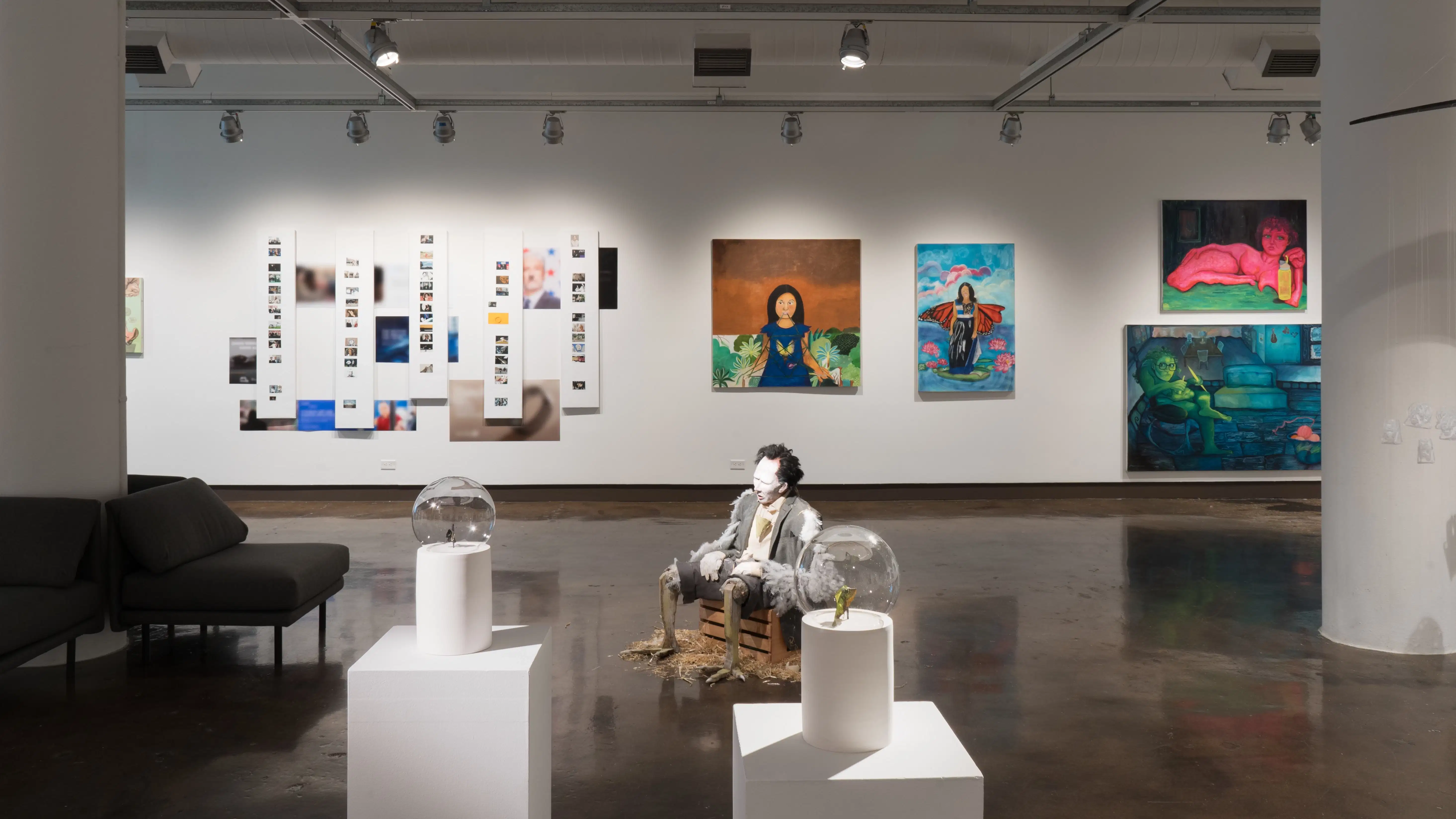 In the foreground, two glass globes on ceramic bases sit on top of pedestals. Each globe contains a wilting leaf. Behind them, a sculpture of a man with feathers and chicken legs slouches in the center of the room. Paintings hang on the back wall.