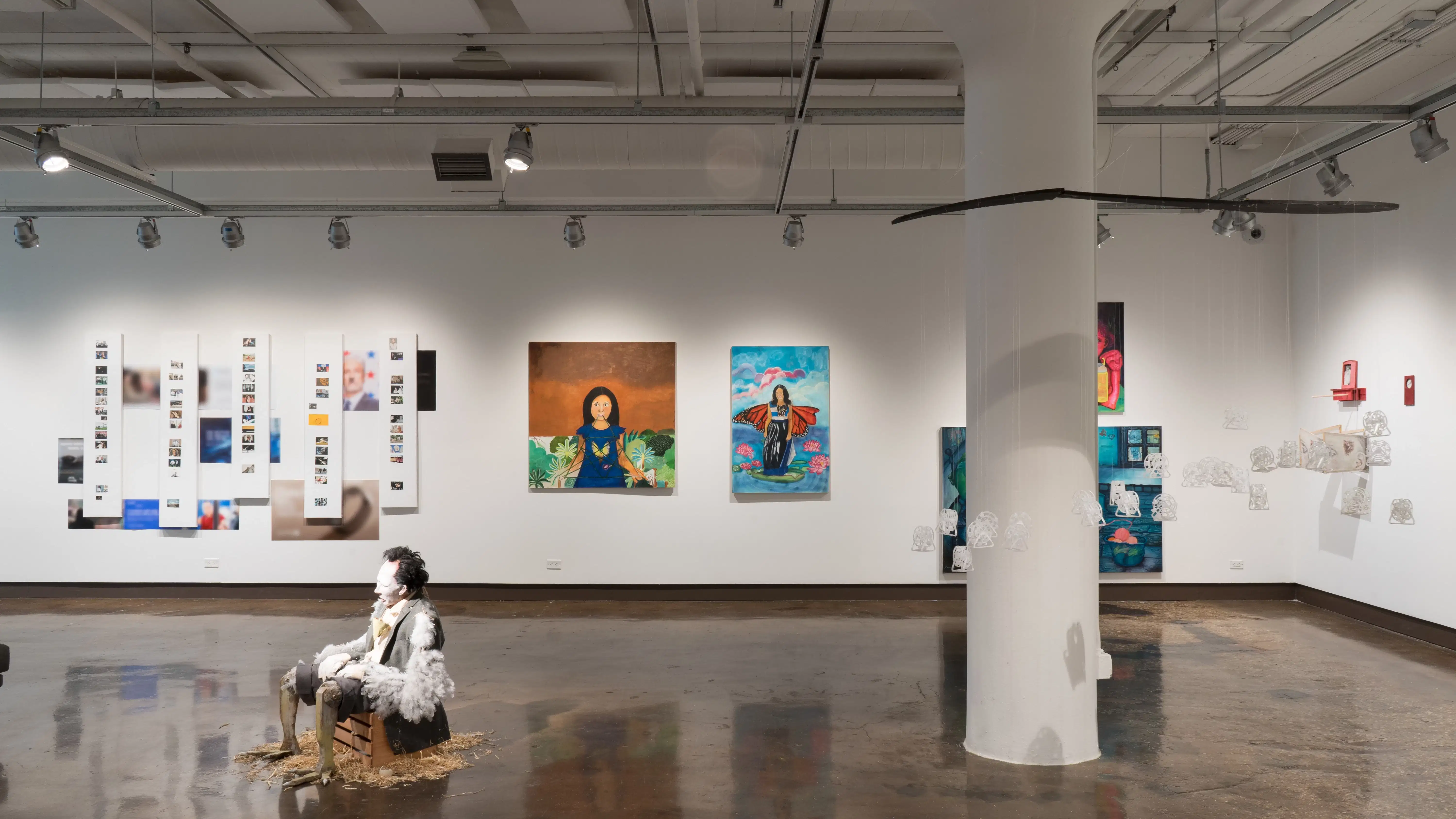 Many paintings hang on the far wall of the gallery. A sculpture of a man with feathers and chicken legs slouches in the center of the room, and to the right, a group of small white plastic sculptures are suspended from the ceiling.