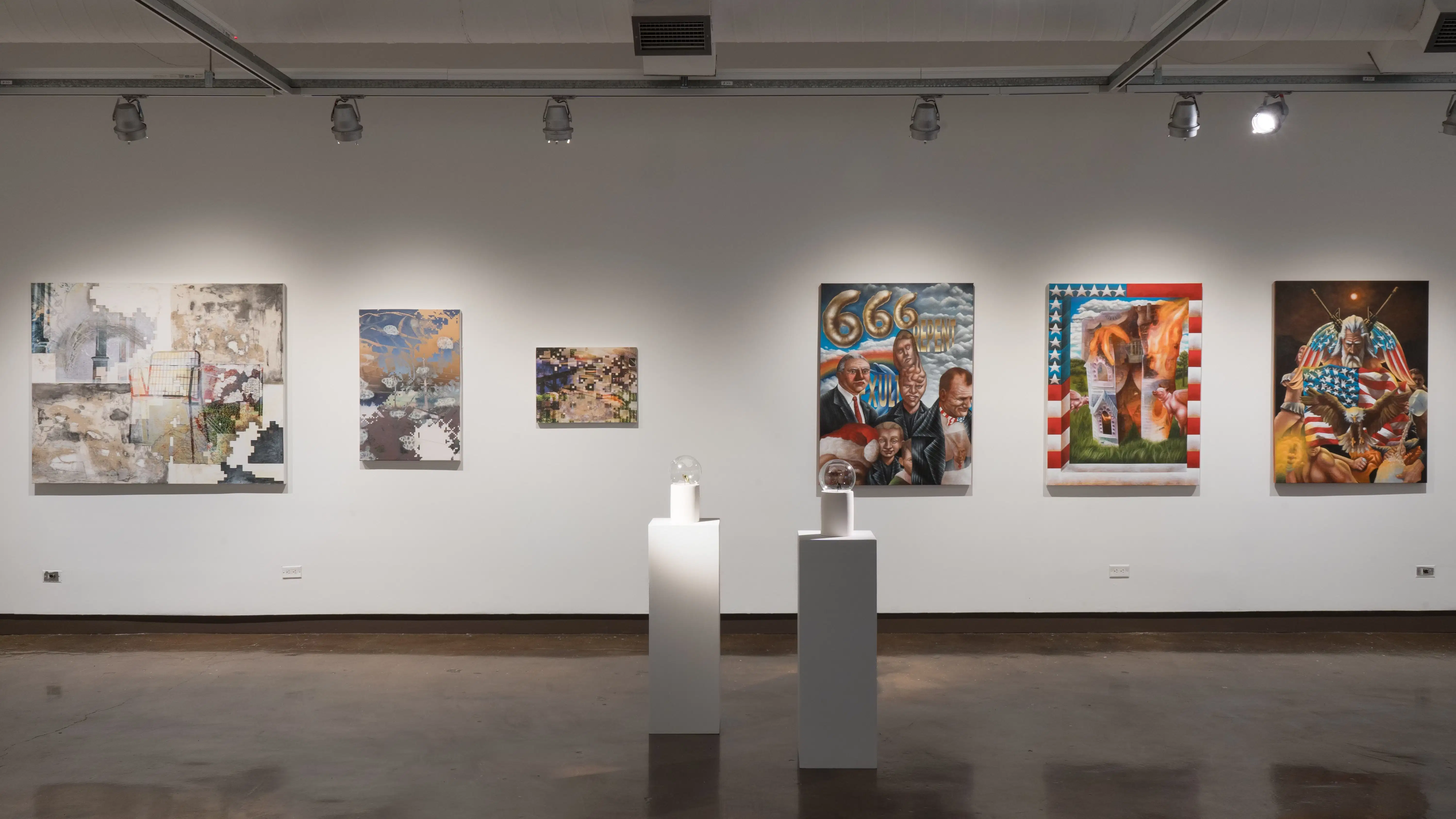 Three mixed media works on canvas hang in a row on one wall. Three paintings depicting imagery related to Christianity, white nationalism, and the United States hang to their right. Two glass globes sit on pedestals in front of them.