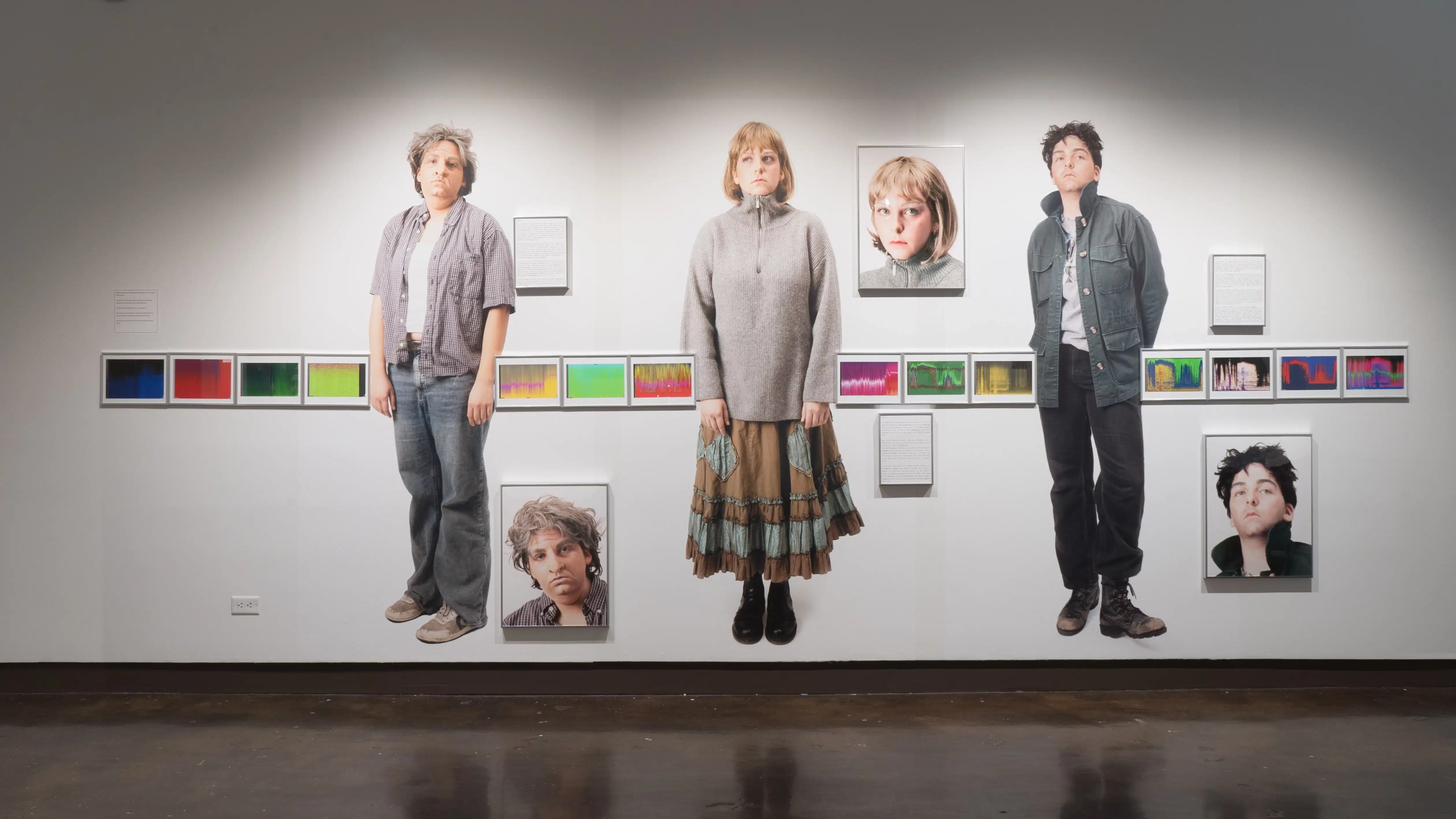 Three larger than life photographs of people in different outfits are displayed on a wall. Smaller framed photographs of their faces hang beside them. A line of framed sound wave images runs horizontally across the installation.