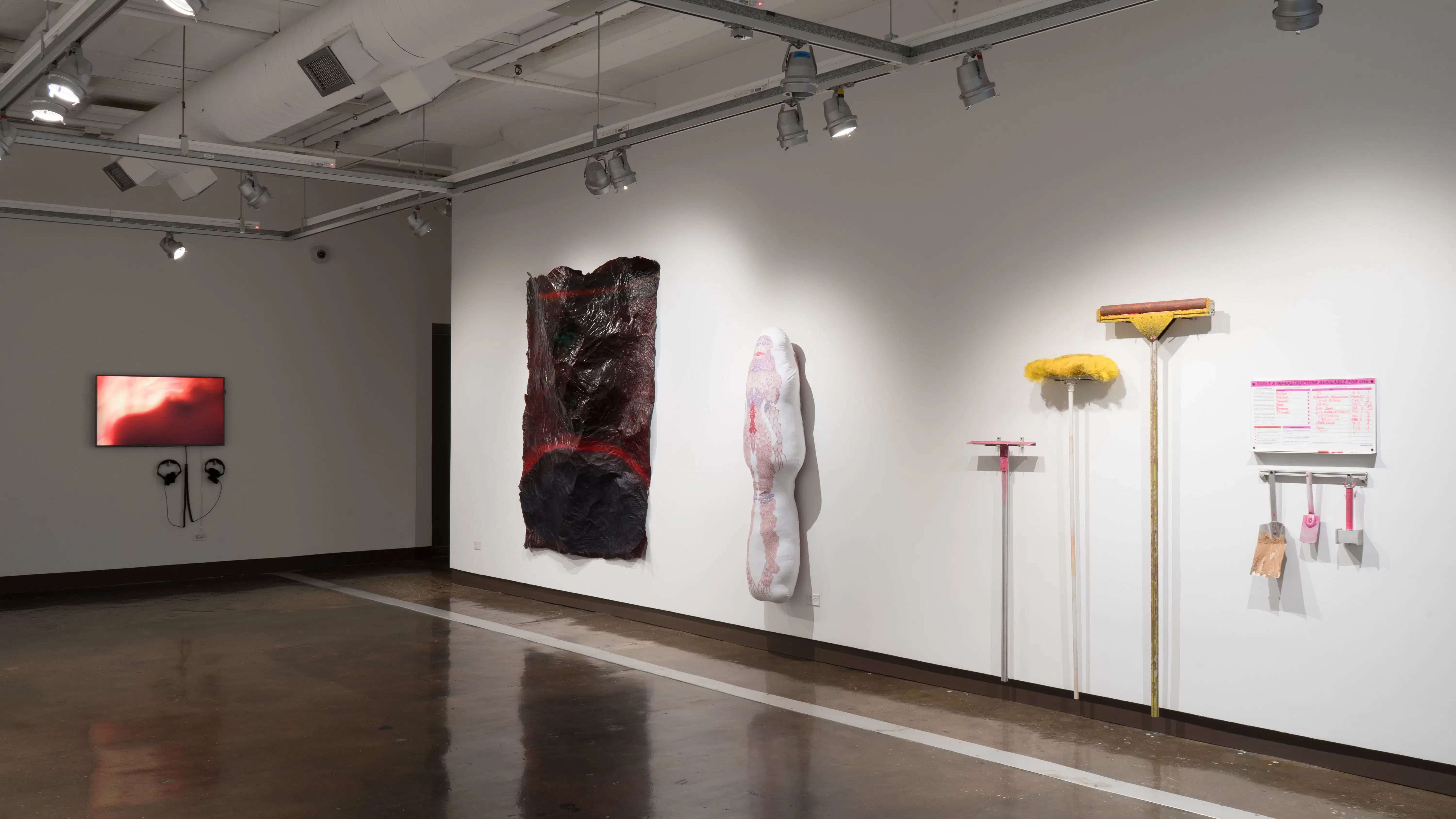 A monitor at the far end of gallery two displays a red, blurry image. On the adjacent wall, a large, sheet-like paper sculpture hangs next to a body pillow-like textile sculpture. Closer, several pink and yellow tools hang with a small dry-erase board.