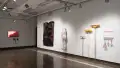 A monitor at the far end of gallery two displays a red, blurry image. On the adjacent wall, a large, sheet-like paper sculpture hangs next to a body pillow-like textile sculpture. Closer, several pink and yellow tools hang with a small dry-erase board.