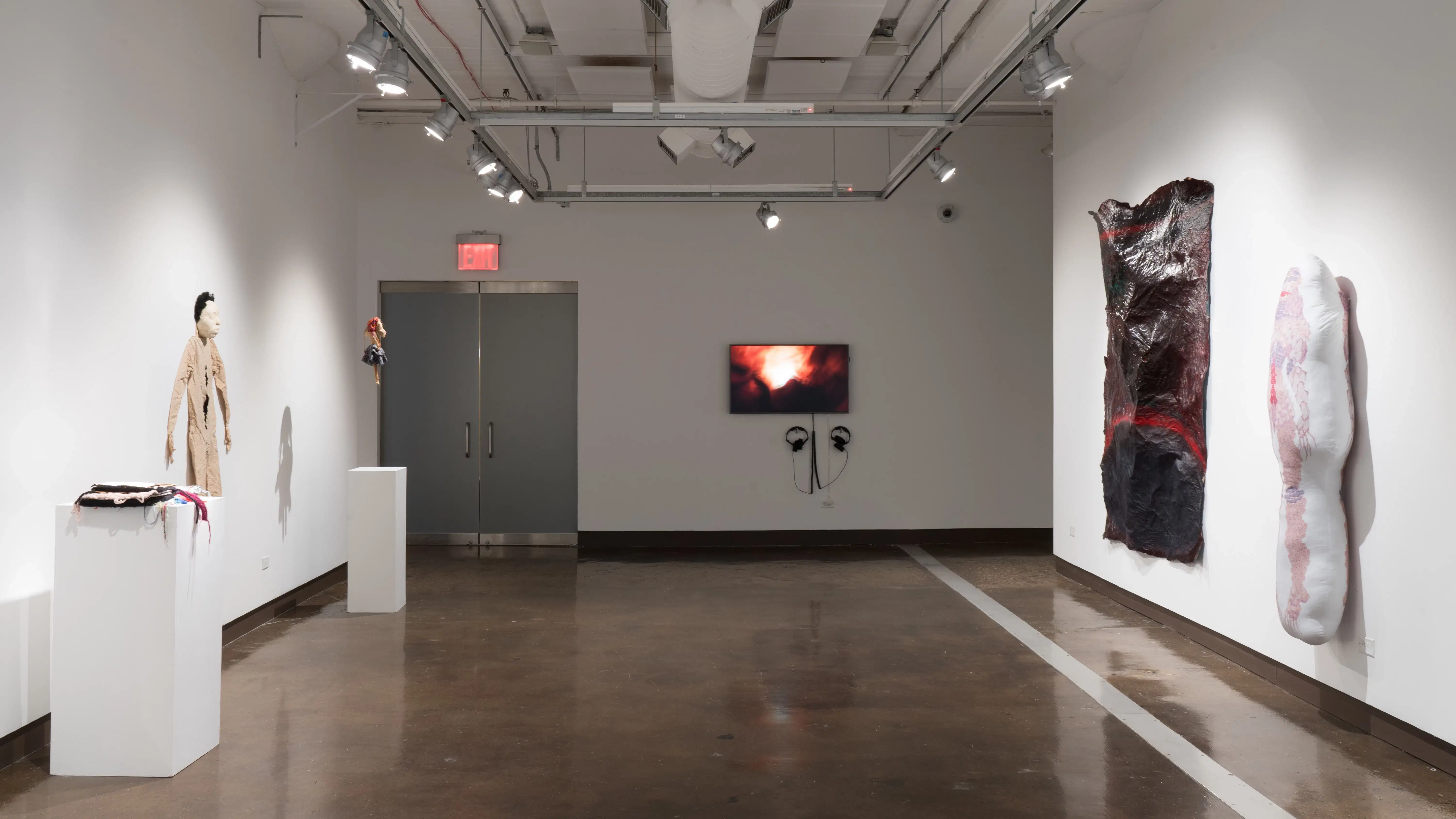 Looking towards the monitor in gallery two, the sheet-like paper sculpture and pillow-like textile sculpture hang on the right wall. To the left, a pedestal holding a textile book sculpture sets beside two figurative sculptures.