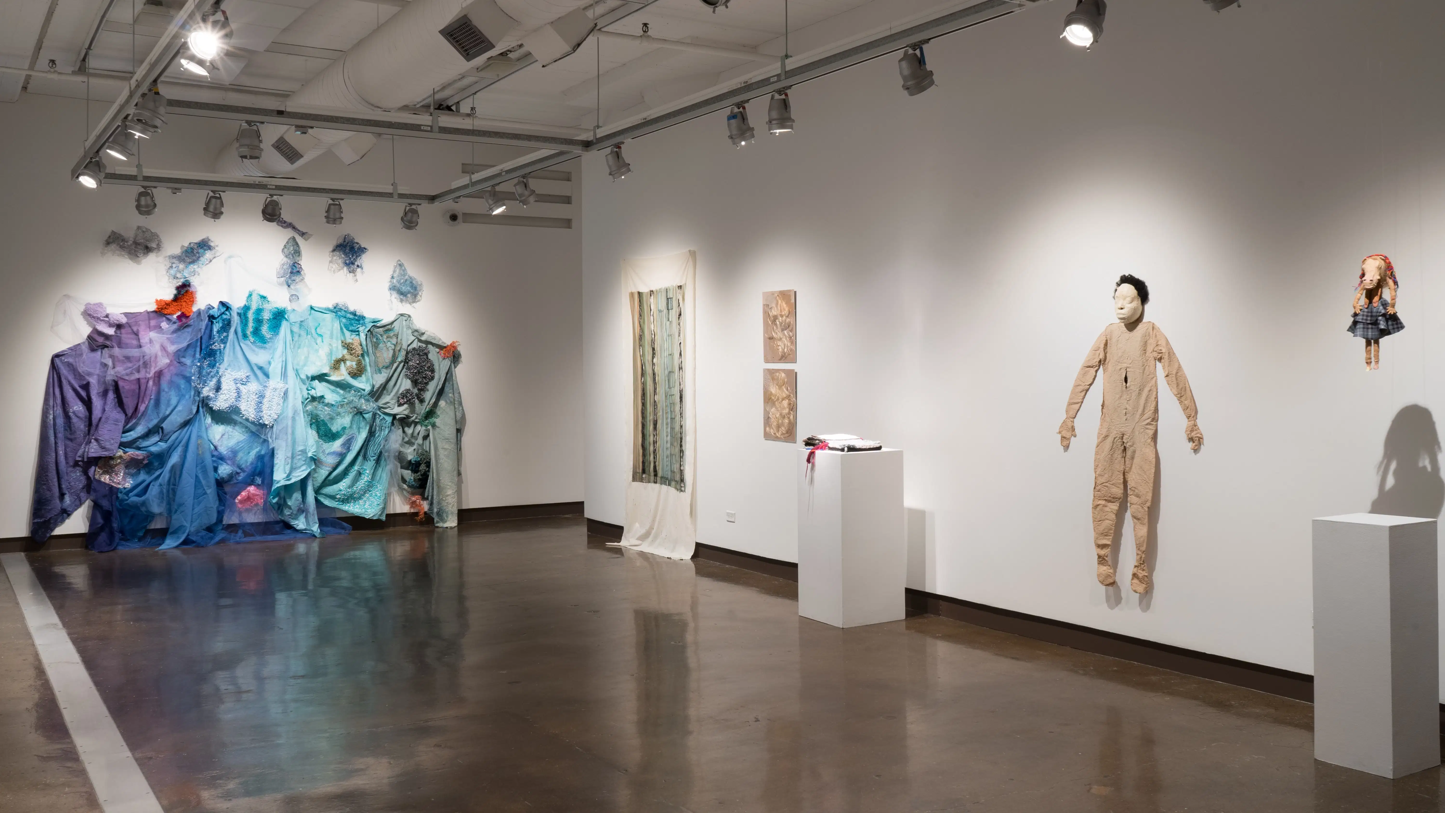 At the other end of gallery two, the wall is filled with a large textile installation. On the adjacent wall, a green painting on loose fabric hangs beside two blonde wigs, squished flat in frames. Closer, a textile sculpture sits on a pedestal next to two figurative sculptures.
