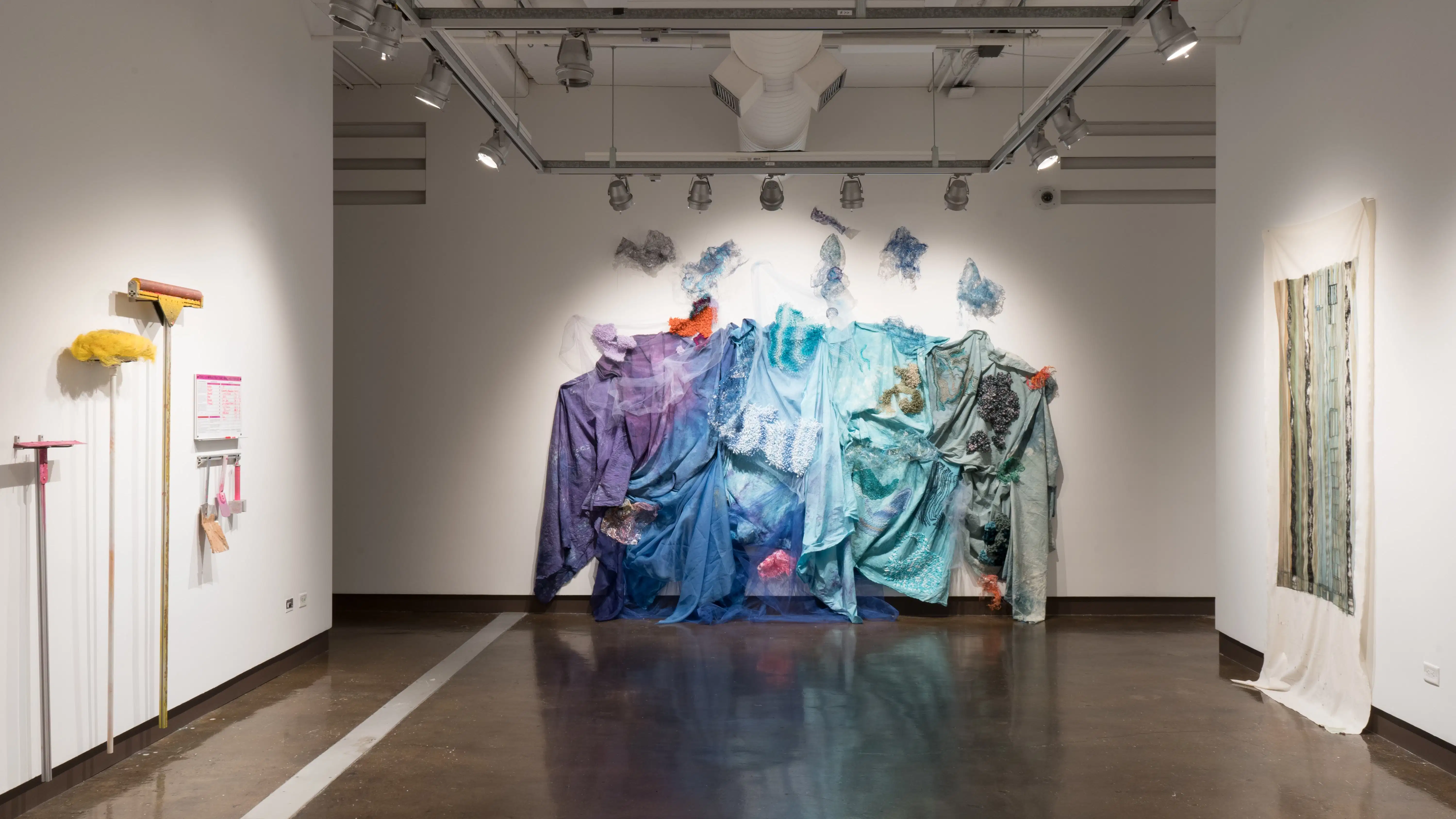 Looking towards the textile installation in gallery two, the painting on loose fabric hangs on the right wall. The pink and yellow tools and dry-erase board hang on the left wall.