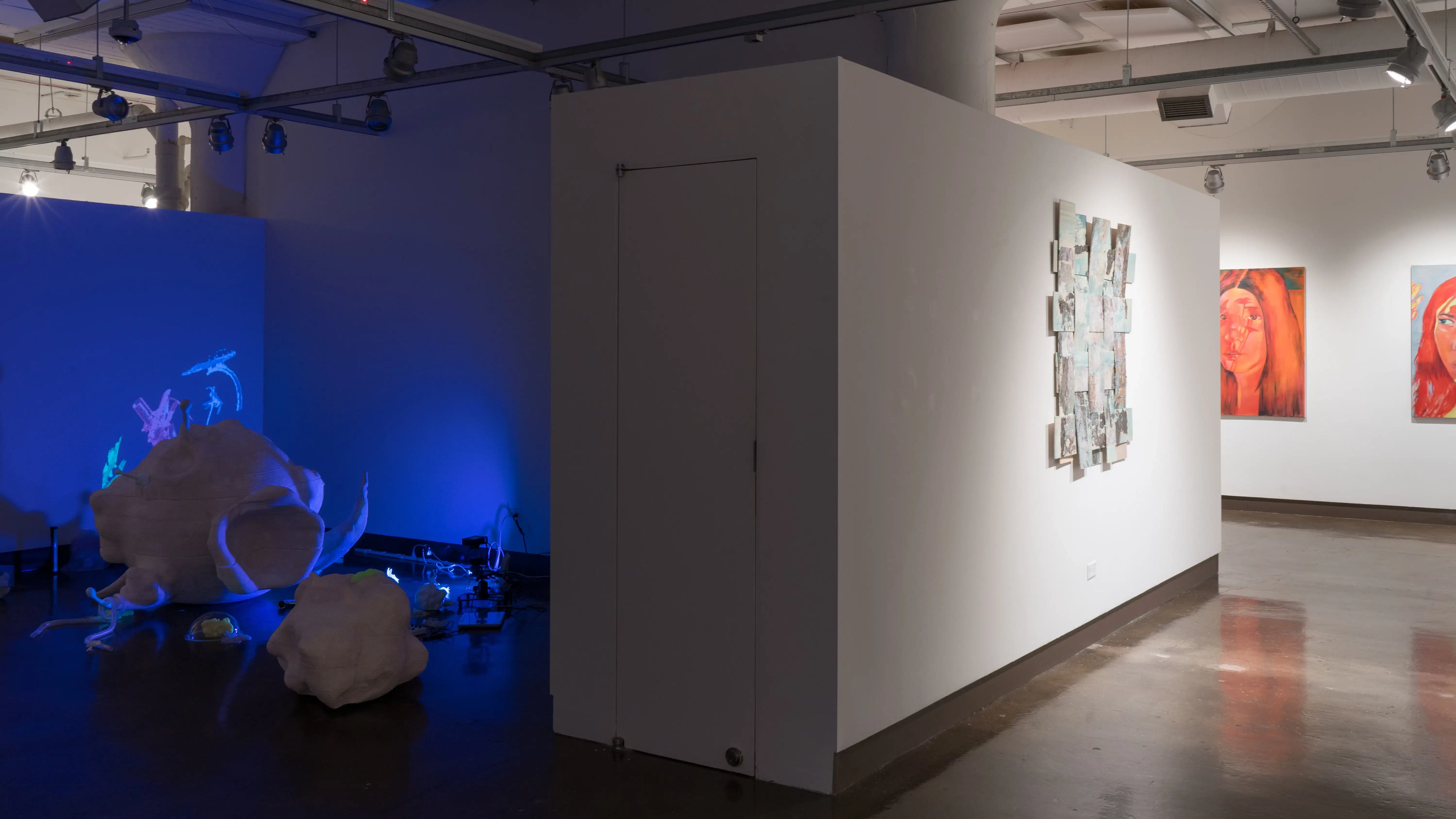 Around the corner, a bluish-purple light glows in the gallery three alcove, among several organically shaped sculptures and objects. A painting on a woven substrate hangs in the right hallway, leading toward gallery four.