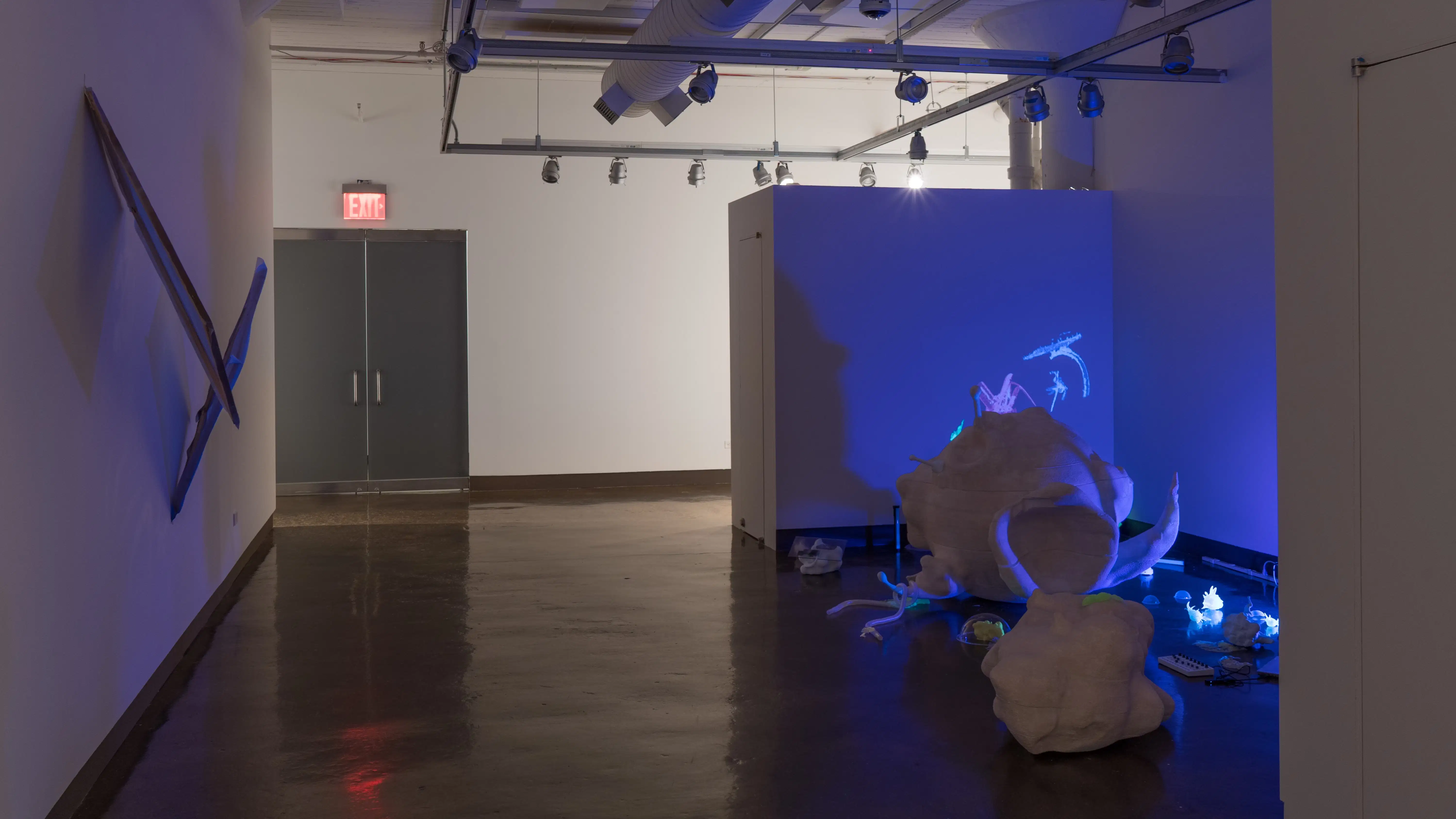 A bluish-purple light glows in the gallery three alcove, among several organically shaped sculptures and objects. Across from the alcove, two stretched paintings lift off of the wall.