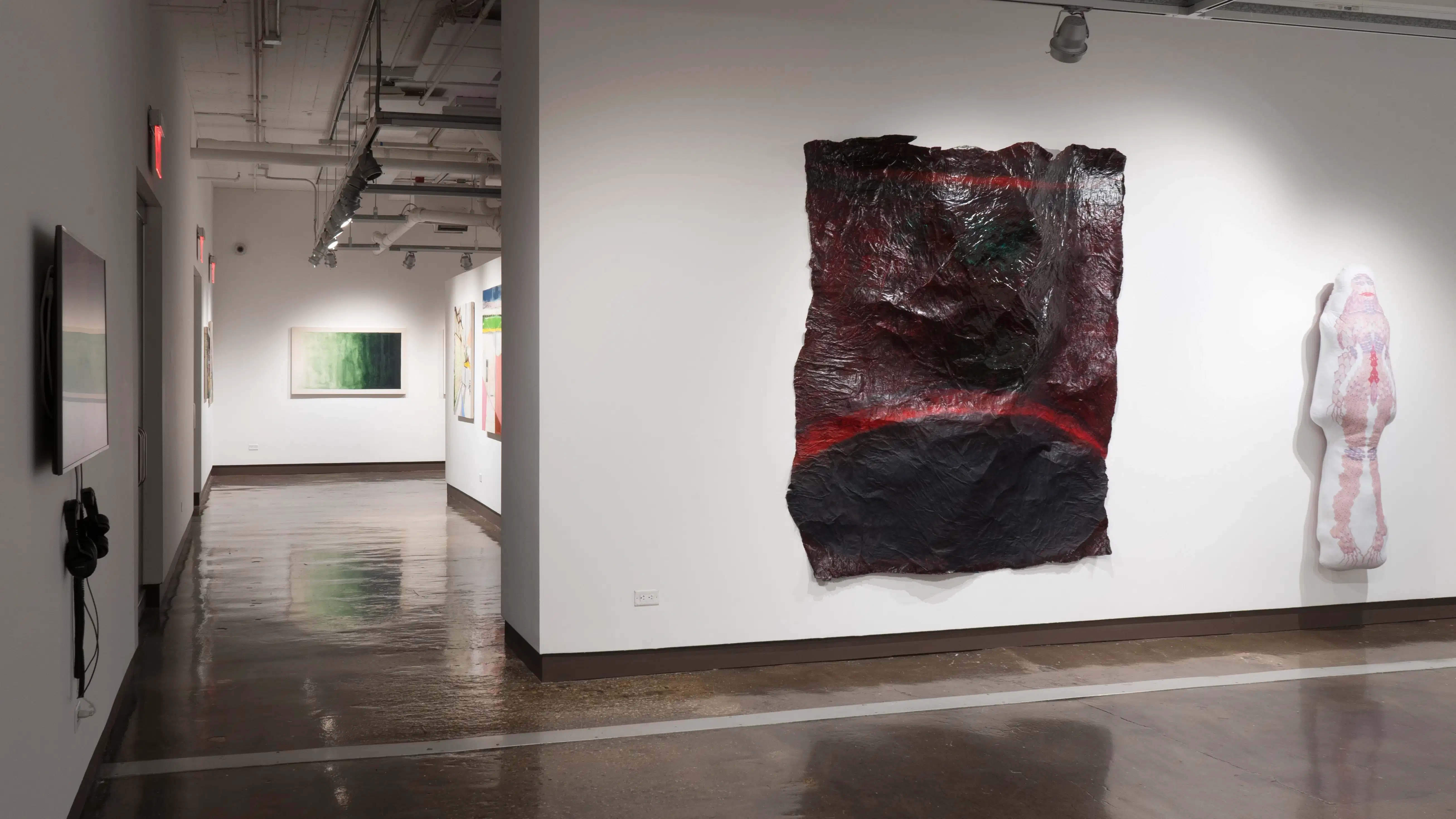 Looking down the left hallway, works in gallery four are visible. Closer, a large sheet-like paper sculpture and a pillow-like textile sculpture hang on the wall in gallery two.