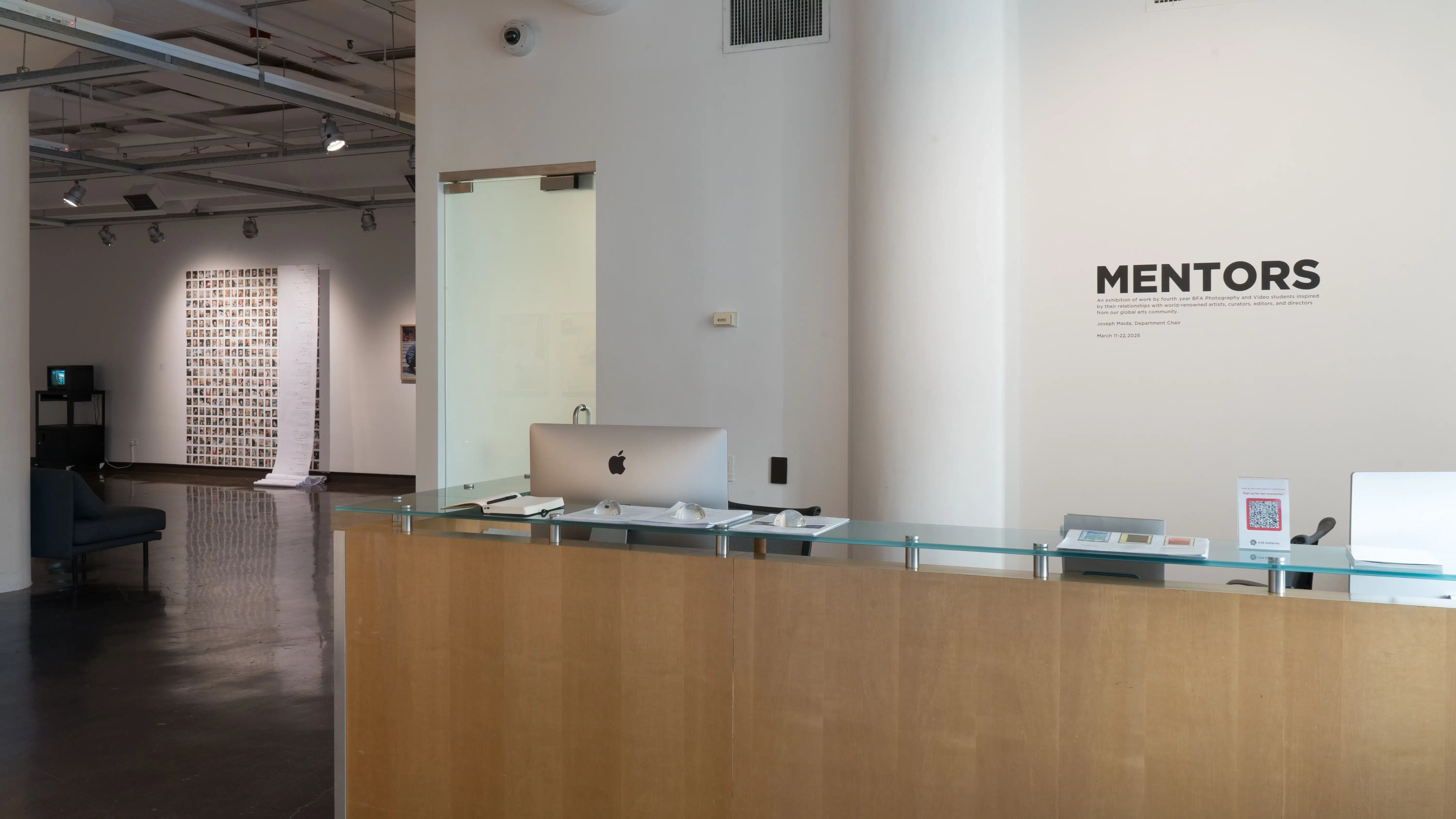 Front desk of gallery with “MENTORS” black decal on wall. Exhibition artworks featured in background. 