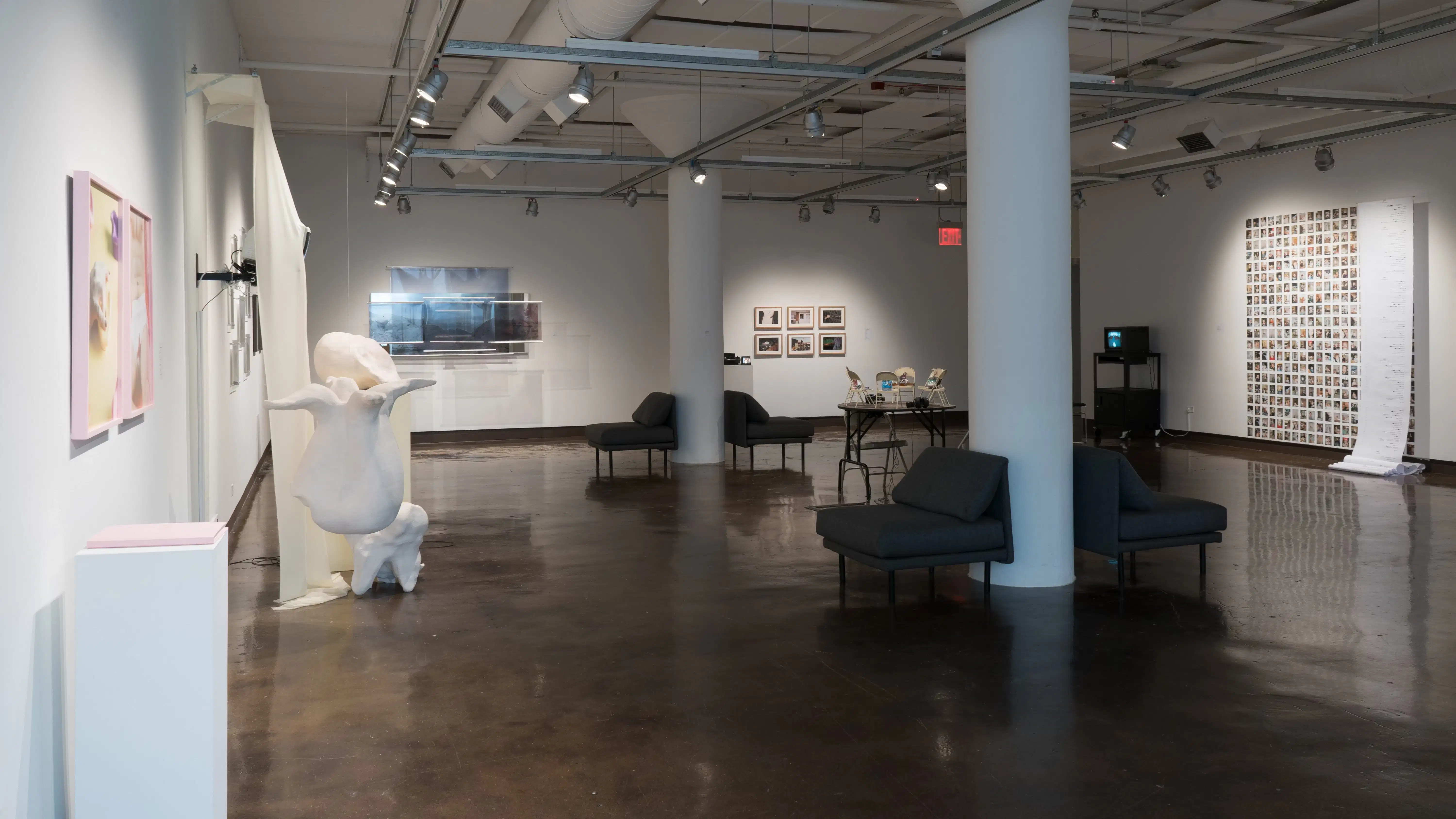 General view of gallery one space, walls lined with print and installation based work, along with a table piece in the center of the room. Couches set facing outwards to the works. 