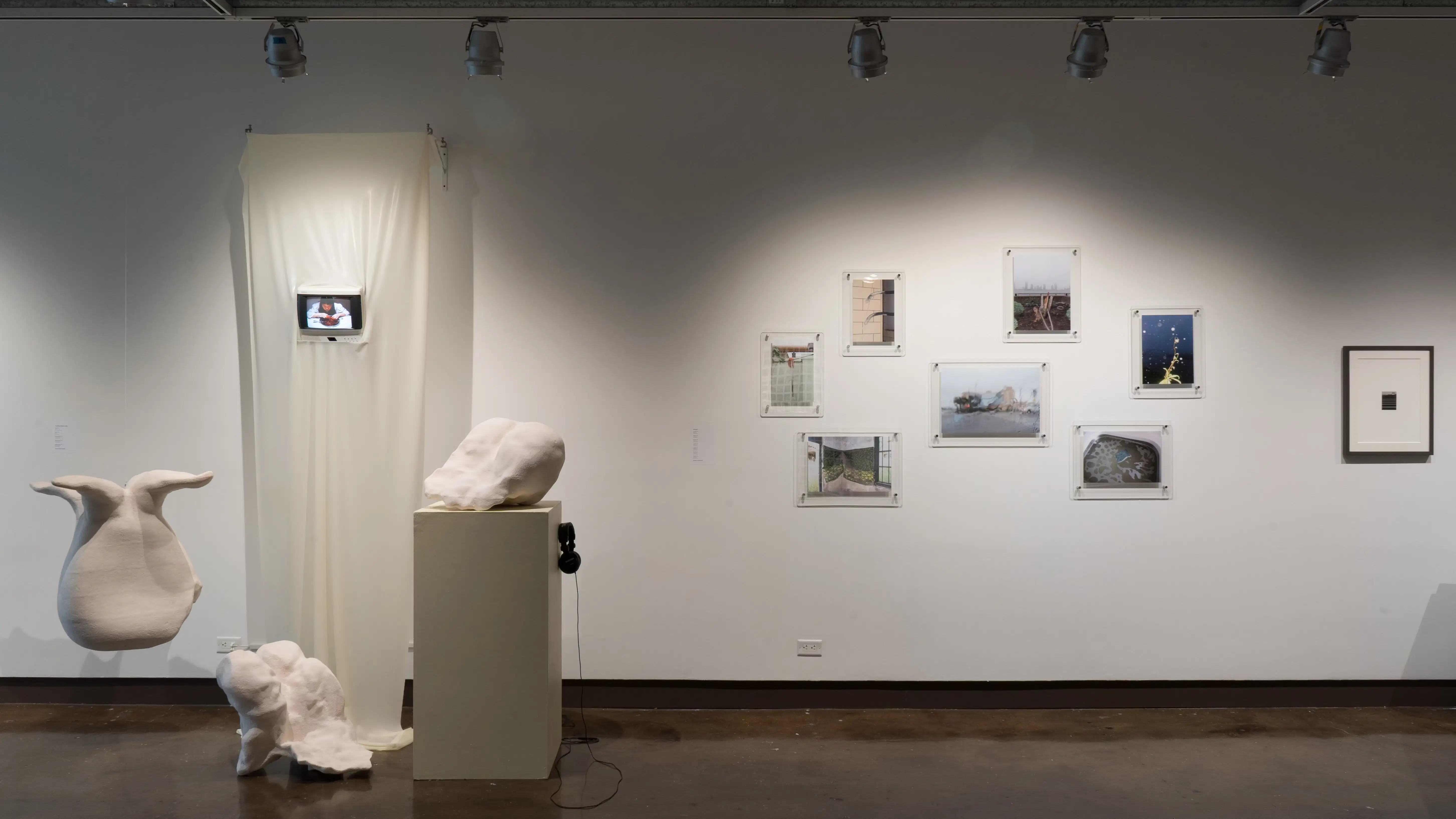 On the left, is a video and sculpture piece, with three off white abstract sculptures. Behind the sculpture is a cream colored latex sheet with a small TV poking through, headphones available on pedestal. On the right, is urban based photography in glass framing. 