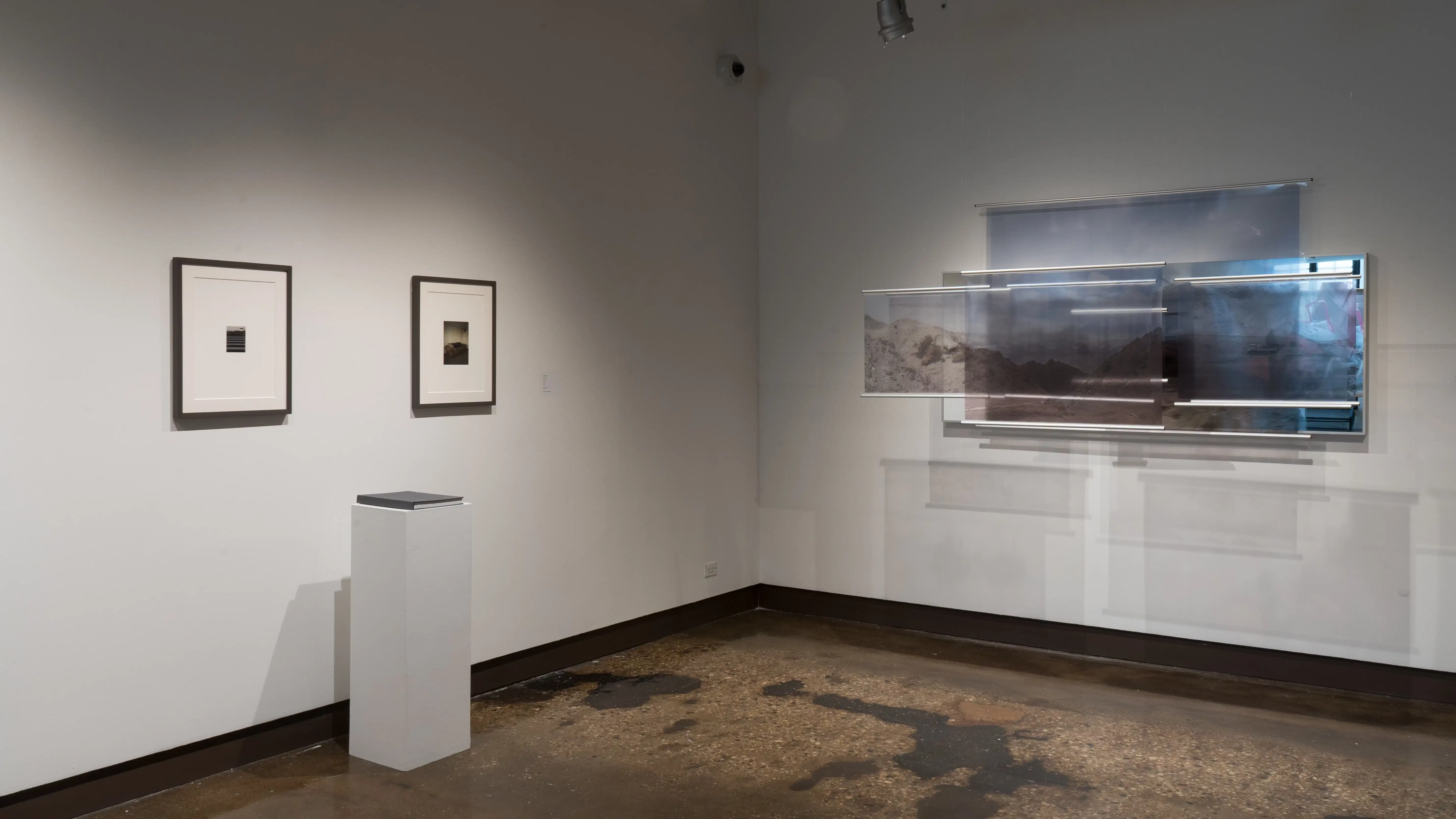Two small scale photography prints in large gray frames with white matte board. In the middle, is a matching gray book on a pedestal. To the right are various transparent photography prints separately layered. A mirror is featured behind photography panels.