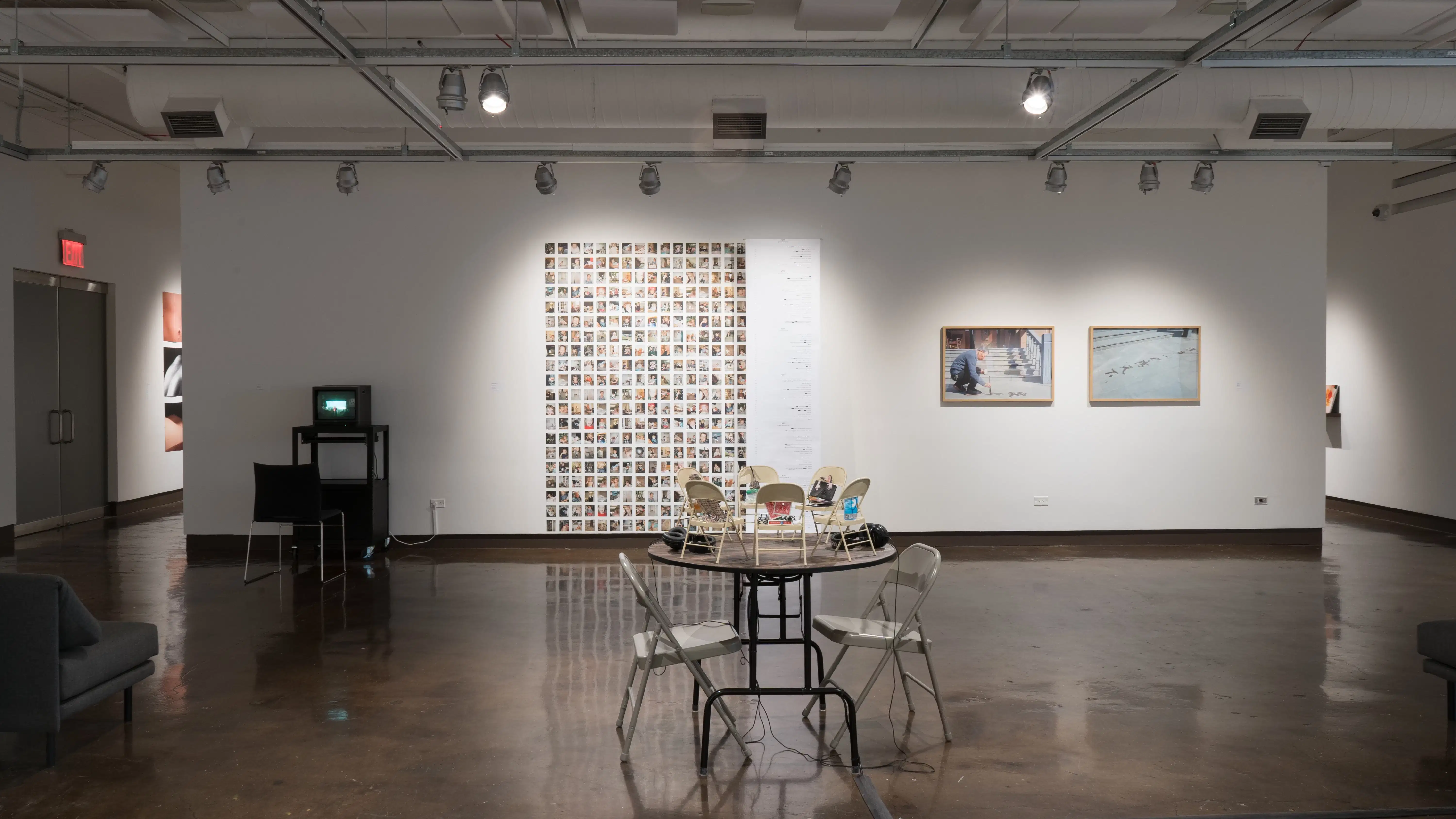 General view of gallery one wall with video piece and print photography. In the foreground is an installation of audio based work on a table with metal chairs on both ends. 