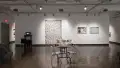 General view of gallery one wall with video piece and print photography. In the foreground is an installation of audio based work on a table with metal chairs on both ends. 