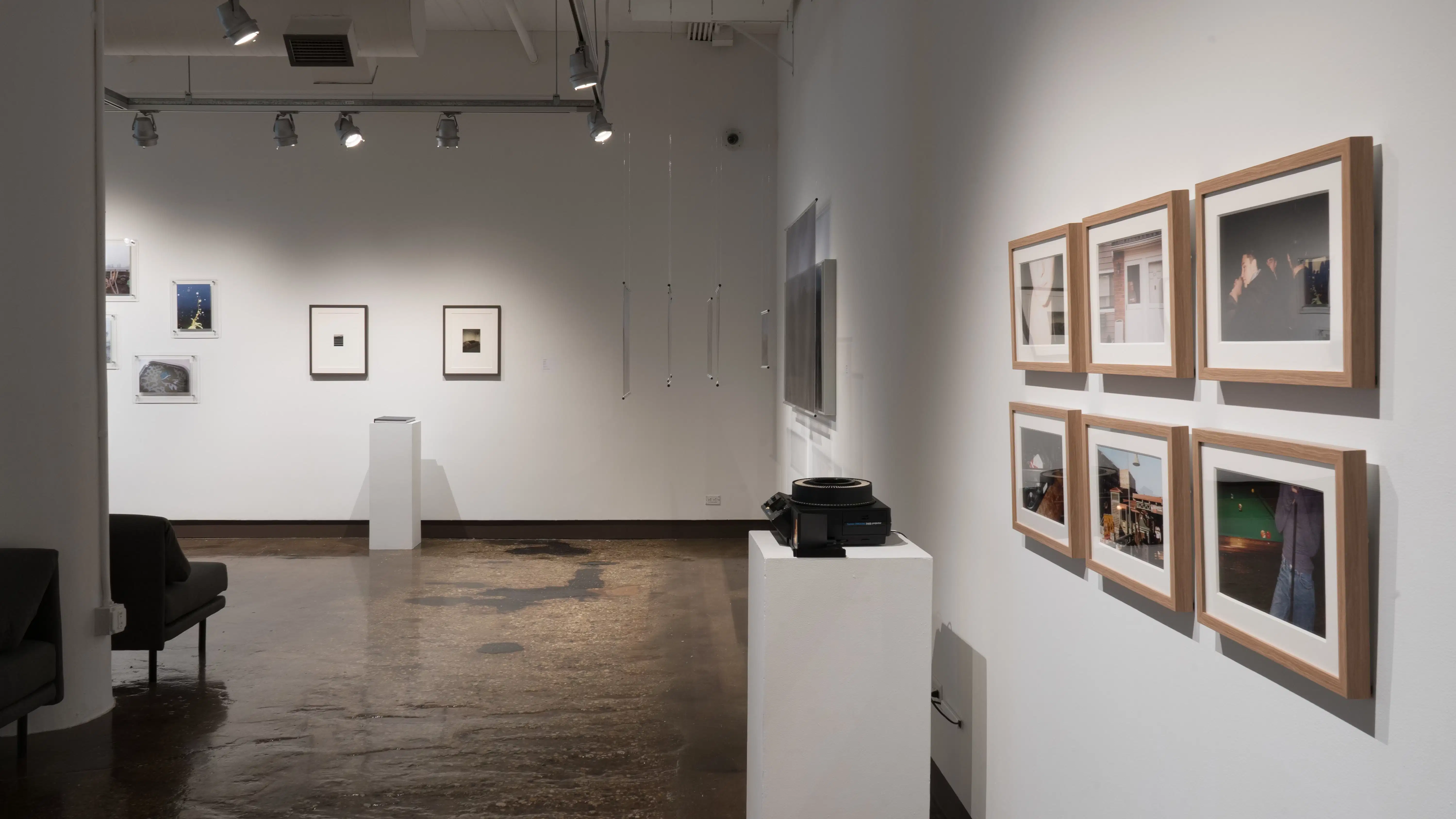 View of gallery one corner with print photography lining the walls, including rotary film display and gray book on pedestals in front of the work. 