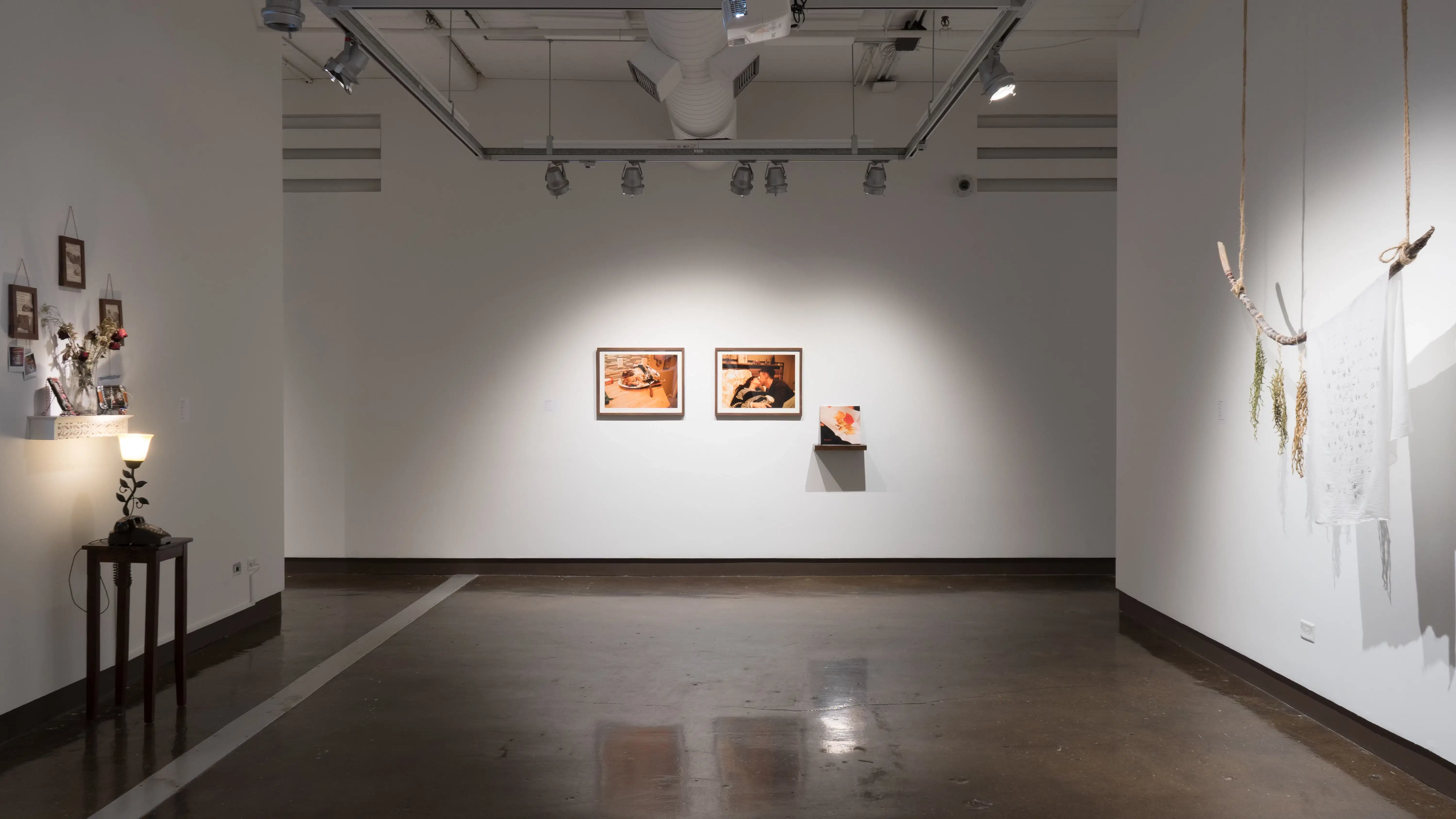 Two large scale warm toned home life photography prints hung in dark wooden frames. A book rests on top of a dark wooden shelf leaning against the wall. On both walls perpendicular to the center piece are three dimensional works. 