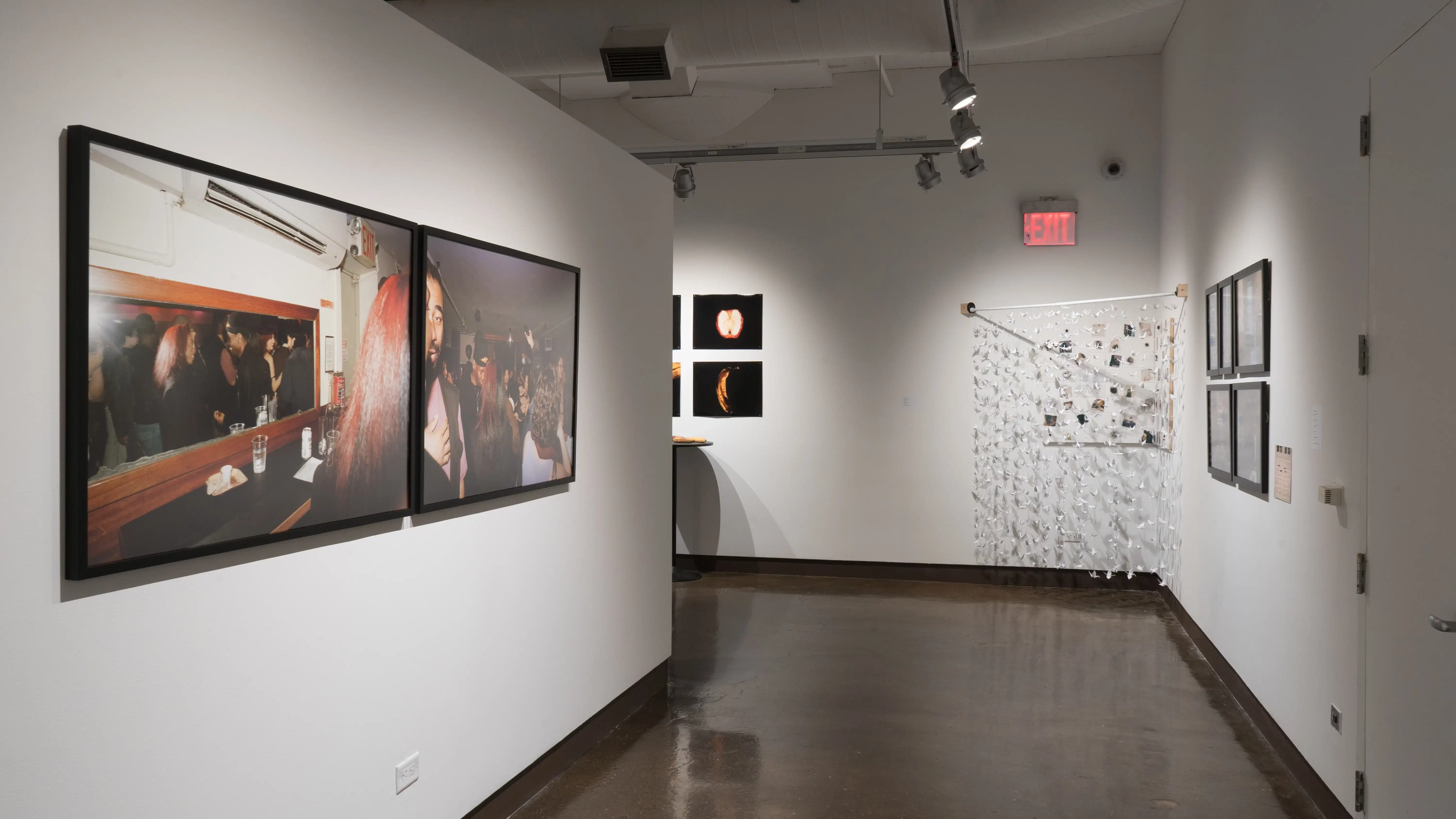 Looking down the gallery hallway featuring nightlife photography on the left. On the right is framed photography, further down in the corner is installation based work with paper cranes hung delicately in front of small scale photographs. 