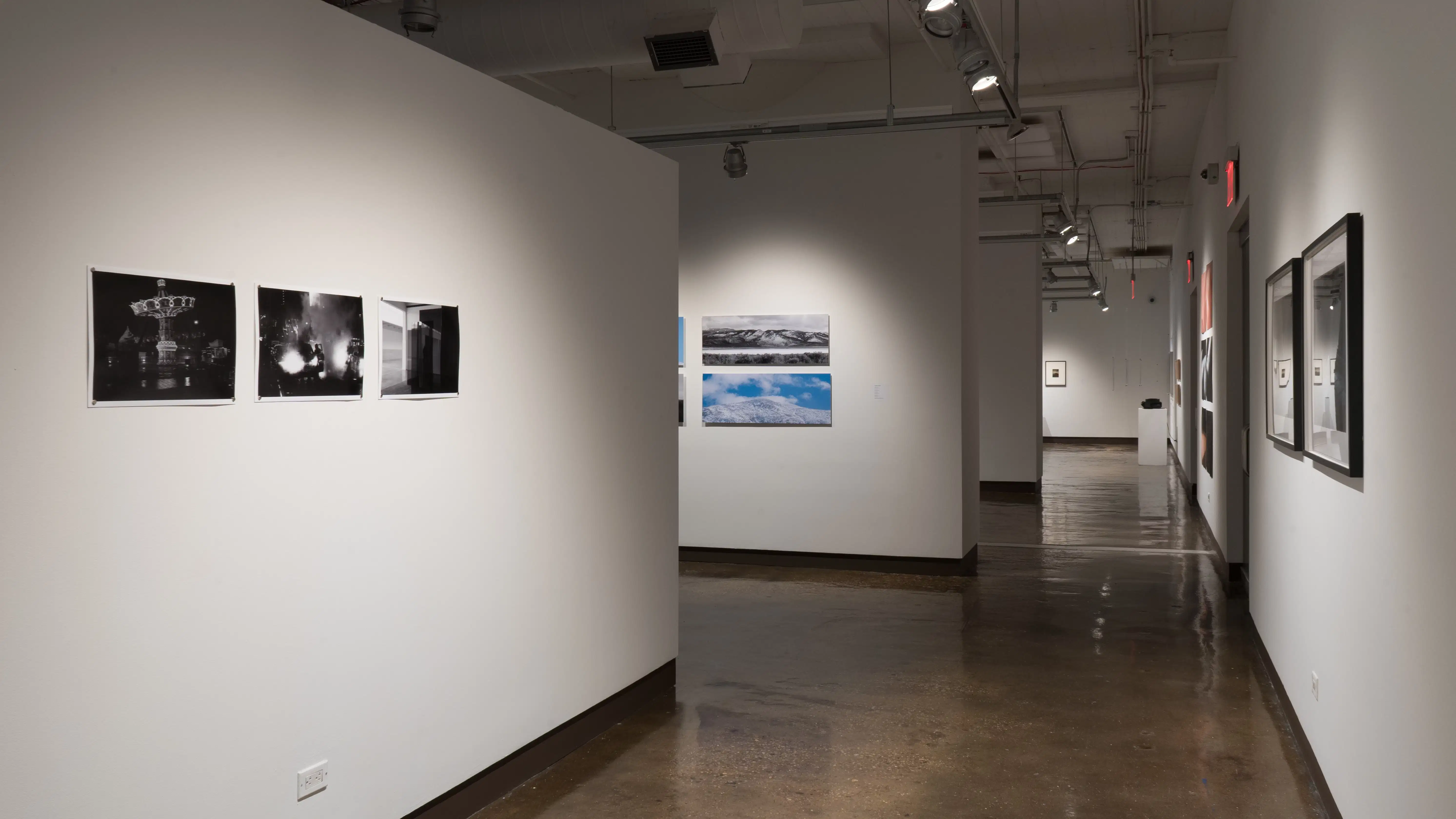Looking down the gallery hallway, three high contrast grayscale photos of the night are pinned to the wall on the left. Further down the right wall are various works of print photography. 
