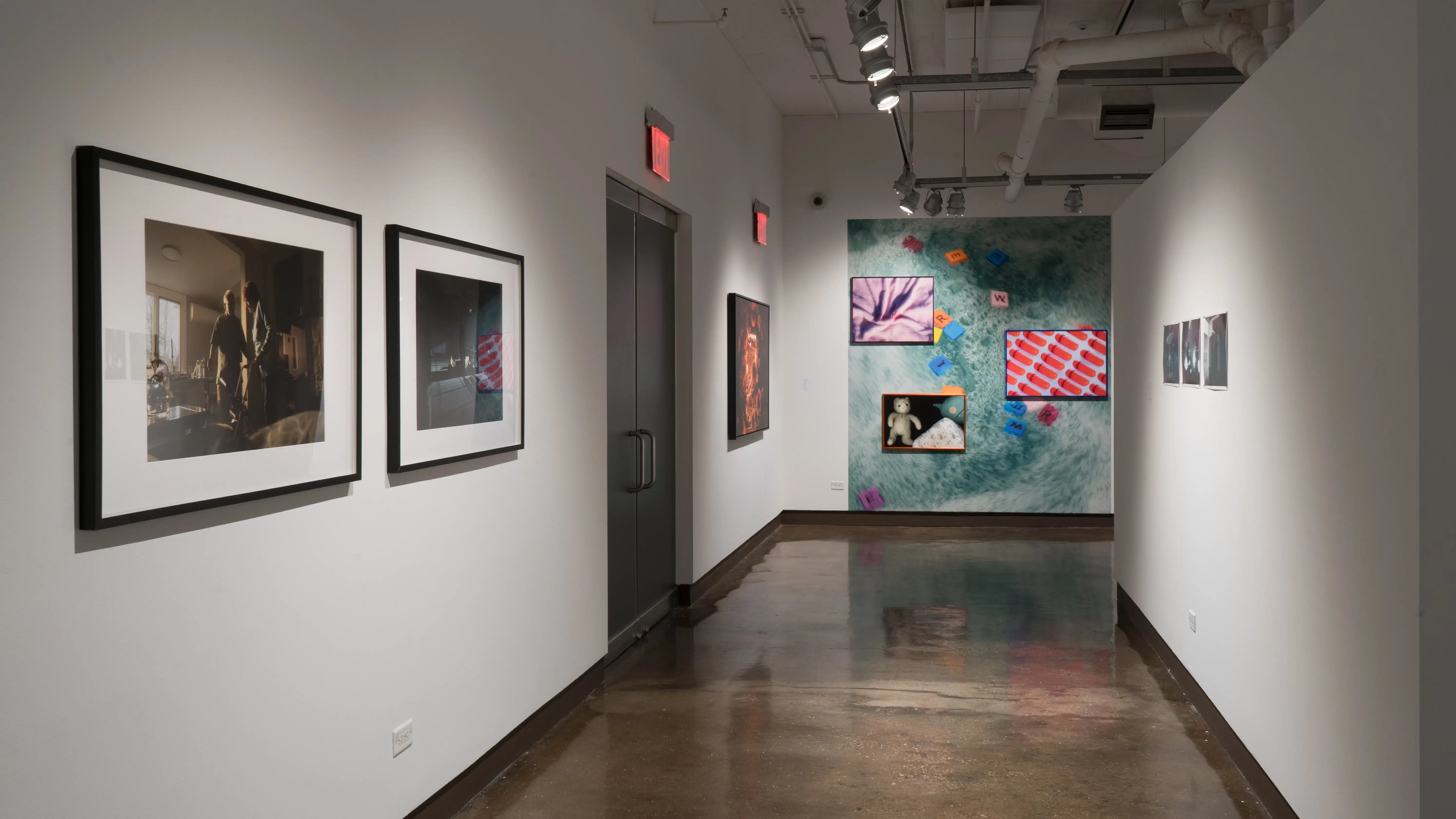 Looking down the gallery hallway, there are various works lining the walls. The wall furthest away has three vibrant print photographs in an alternating sequence with a large scale motion blurred blue background. 