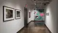 Looking down the gallery hallway, there are various works lining the walls. The wall furthest away has three vibrant print photographs in an alternating sequence with a large scale motion blurred blue background. 