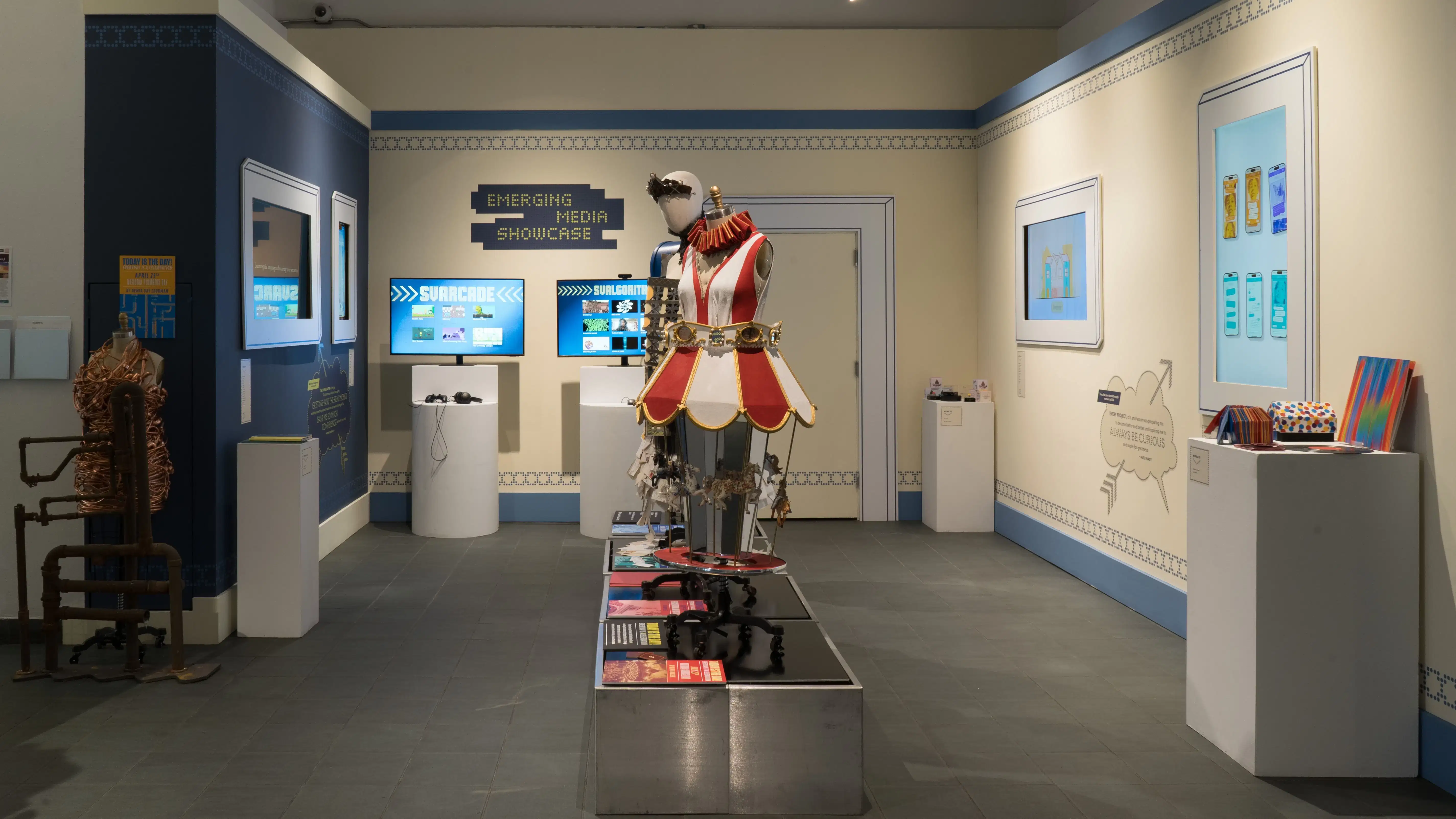 In the lobby of the SVA Gramercy Gallery, a mannequin wearing a carousel costume stands on a metallic pedestal. On the surrounding walls, framed screens play videos of advertising and design projects.