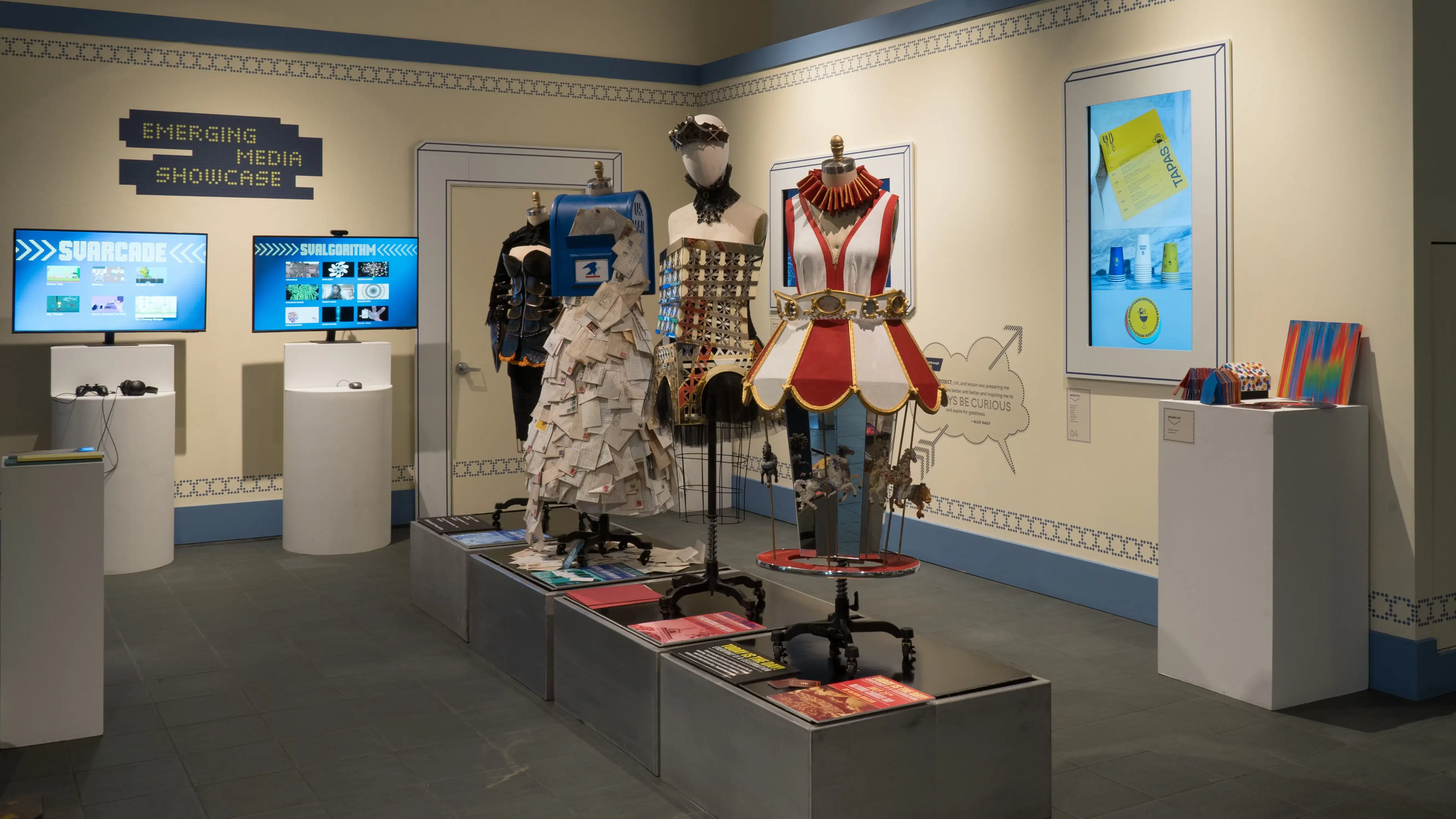 In the lobby of the SVA Gramercy Gallery, four mannequins wearing different costumes stand on a metallic pedestal. On the surrounding walls, framed screens play videos of advertising and design projects.