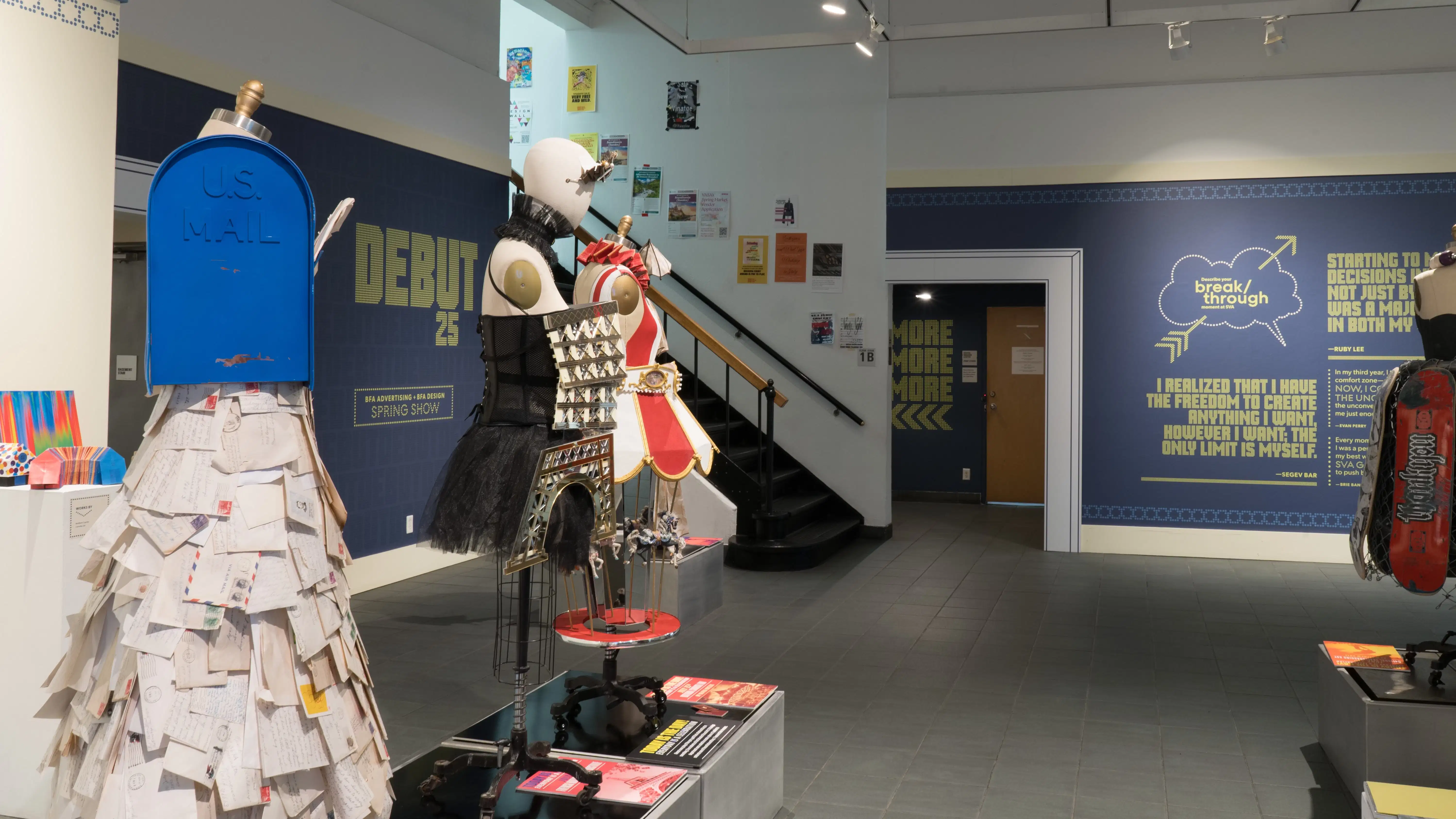 Mannequins dressed in costumes inspired by objects are on display in the center of the room. One costume appears to be a mailbox overflowing with letters; another is a dress made of skateboards and grip tape.