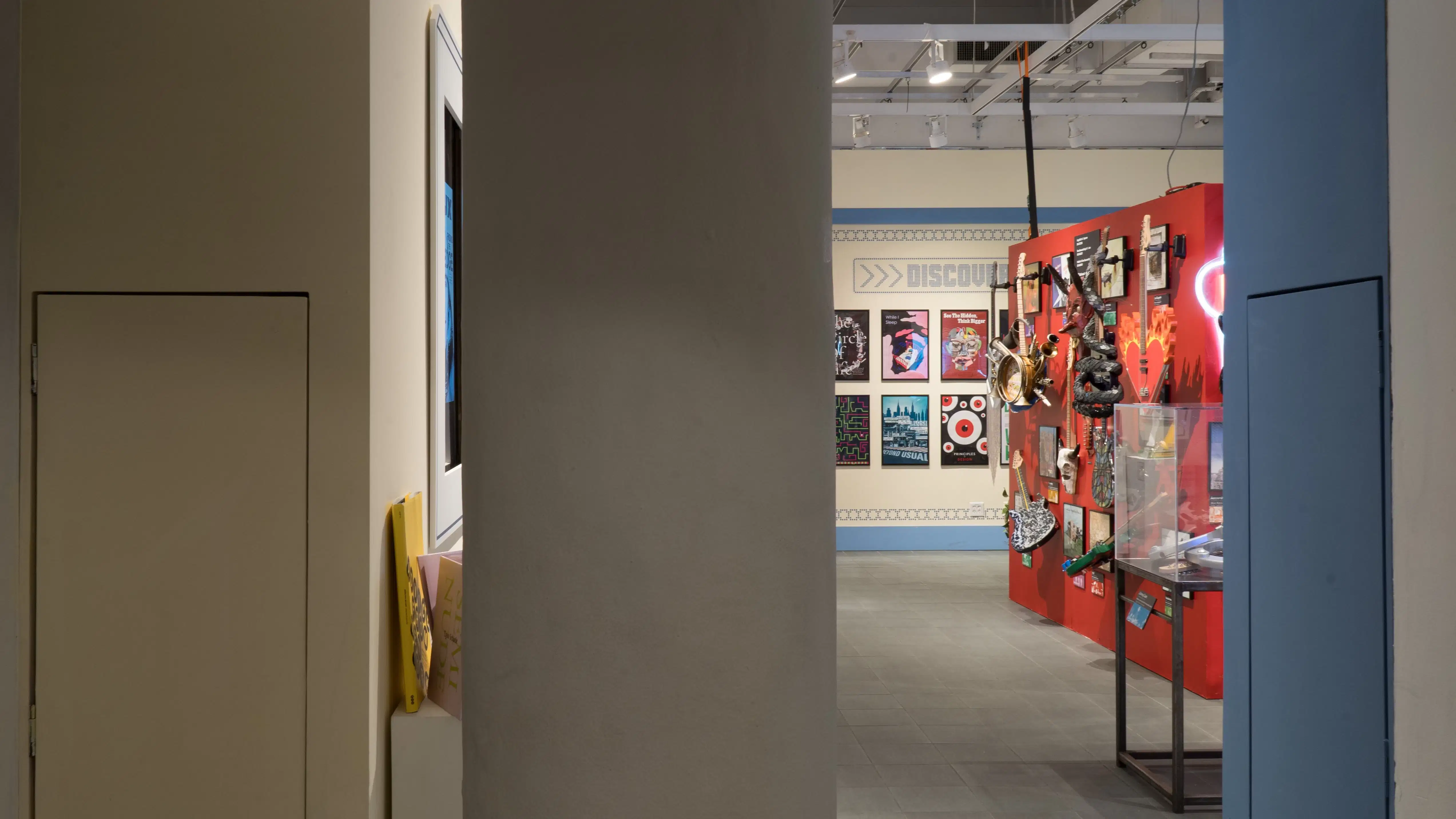 Walking into the SVA Gramercy Gallery back room, a red wall covered in guitars and a yellow wall covered in posters are visible past a column.