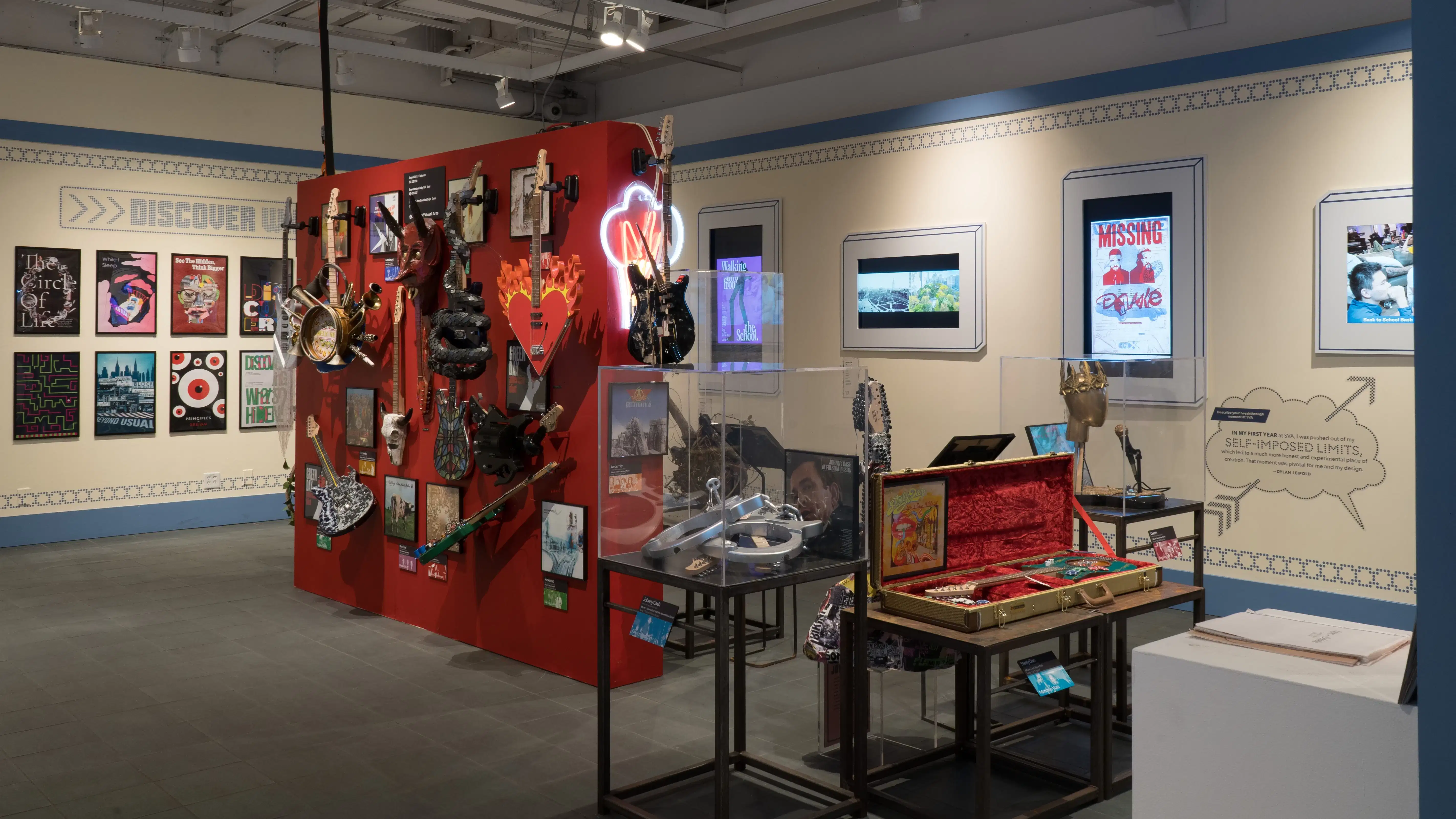 In the back room of the gallery, a red wall in the center of the room displays many guitars with various designs for their bodies. On the surrounding walls, design and advertising projects play on monitors. More sculptural objects are visible on pedestals.