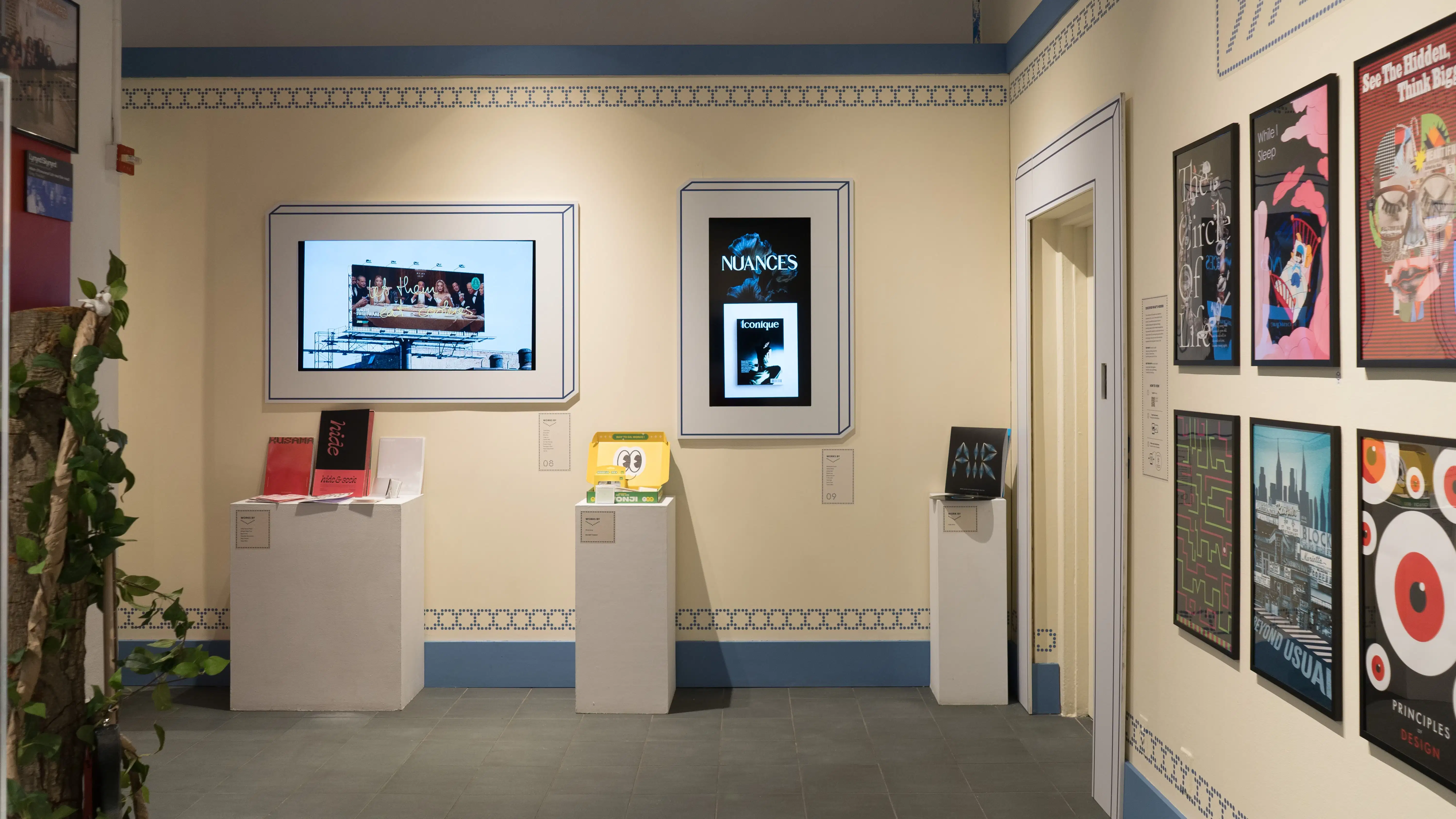 Looking across the gallery, two monitors on a wall display projects while pedestals in front of them hold objects or books. On the perpendicular wall, framed posters are hung in two rows.