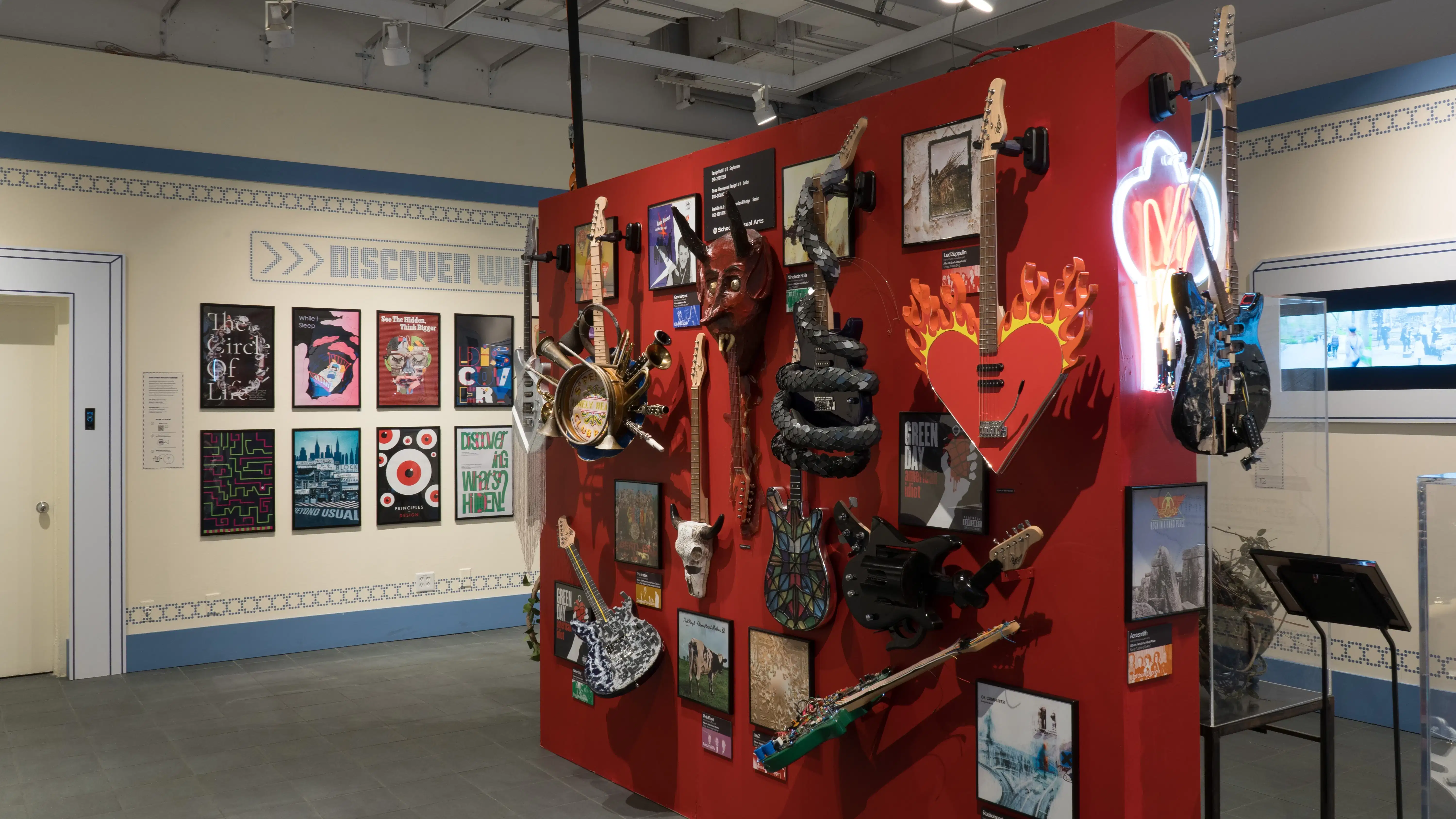 A red wall in the center of the room displays many guitars with various designs for their bodies, like snakes and stained glass windows. On the surrounding walls, design and advertising projects play on monitors.