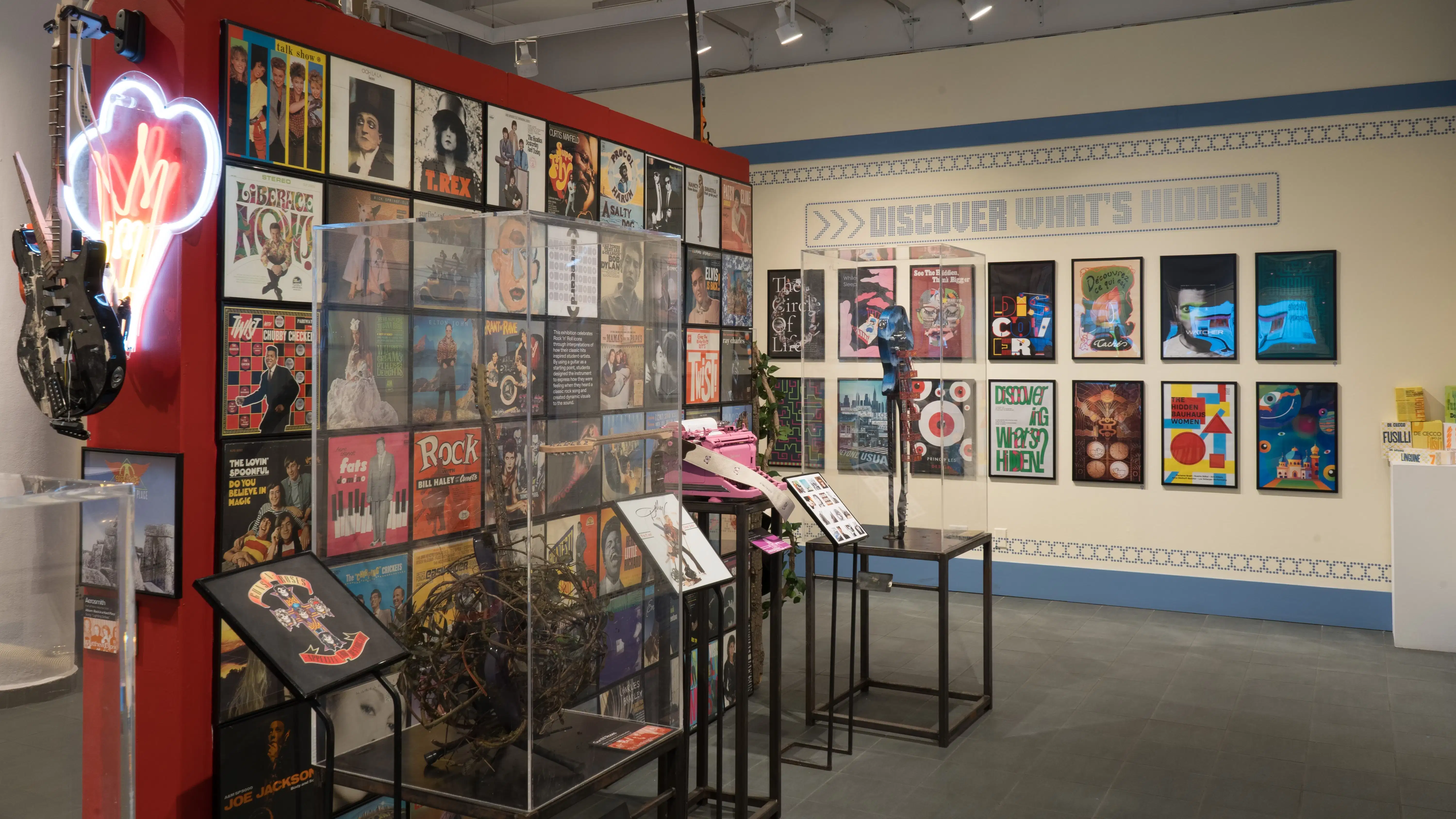 On the opposite side of the red wall, more guitars are presented on pedestals covered by plexiglass bonnets. On the wall beyond them, framed posters are organized into two horizontal rows.