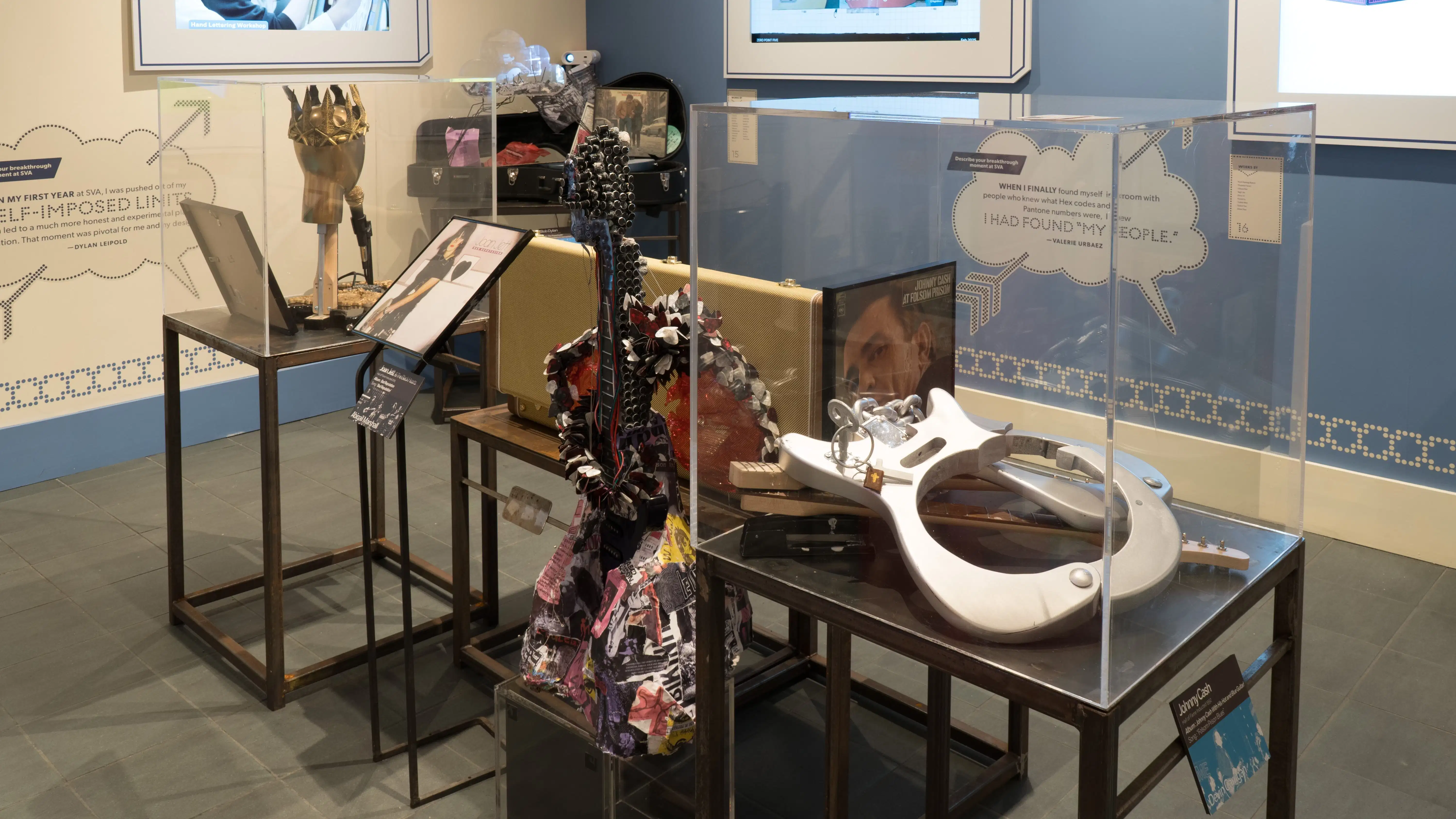 More unique guitars are displayed on pedestals grouped on one side of the room. Beyond them, design and advertising projects play on monitors.