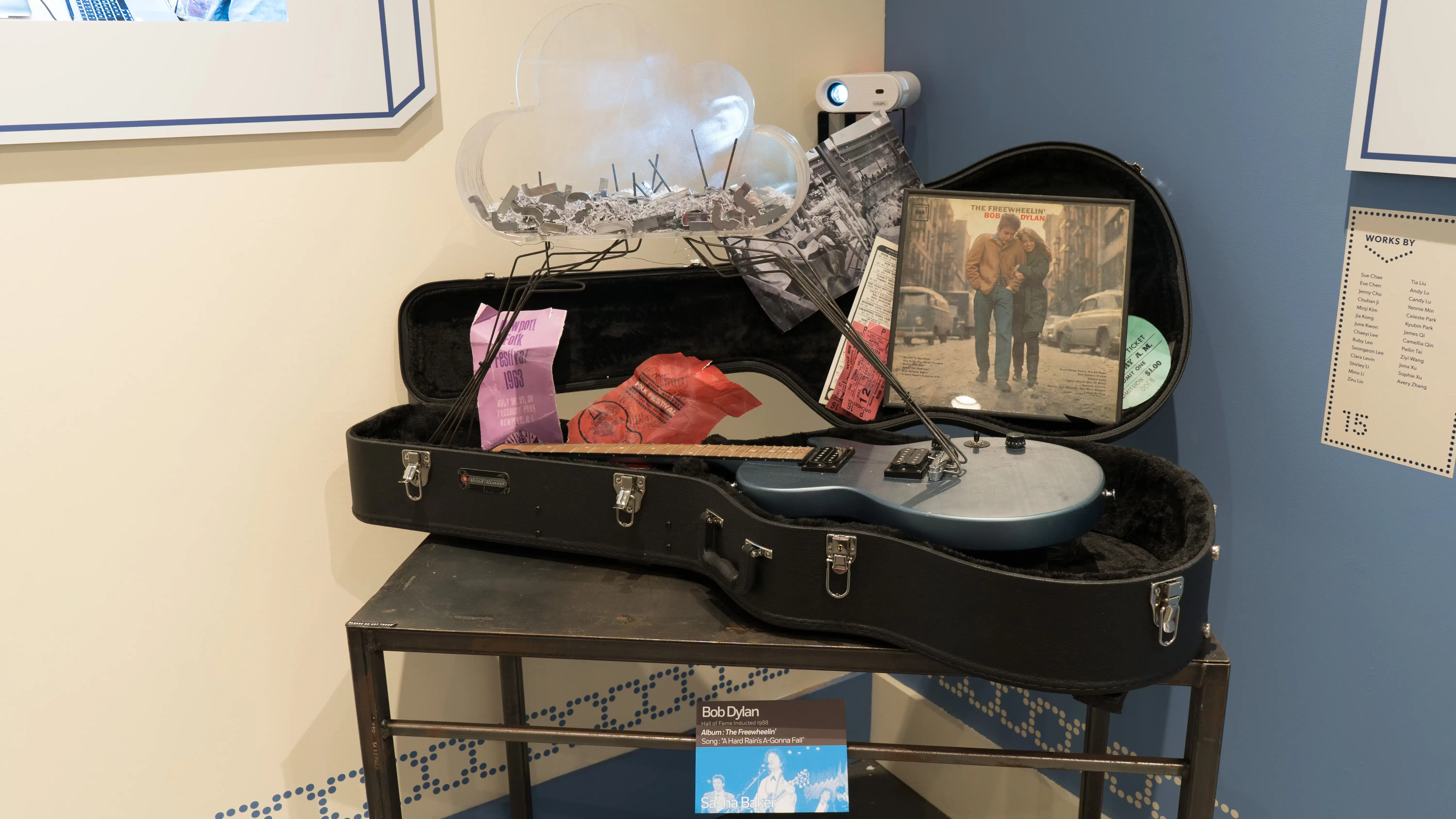 In one corner of the gallery, a grey-blue guitar sits in an open guitar case. Attached to it is a clear plastic cloud filled with bits of paper. A Bob Dylan album leans against the guitar case.