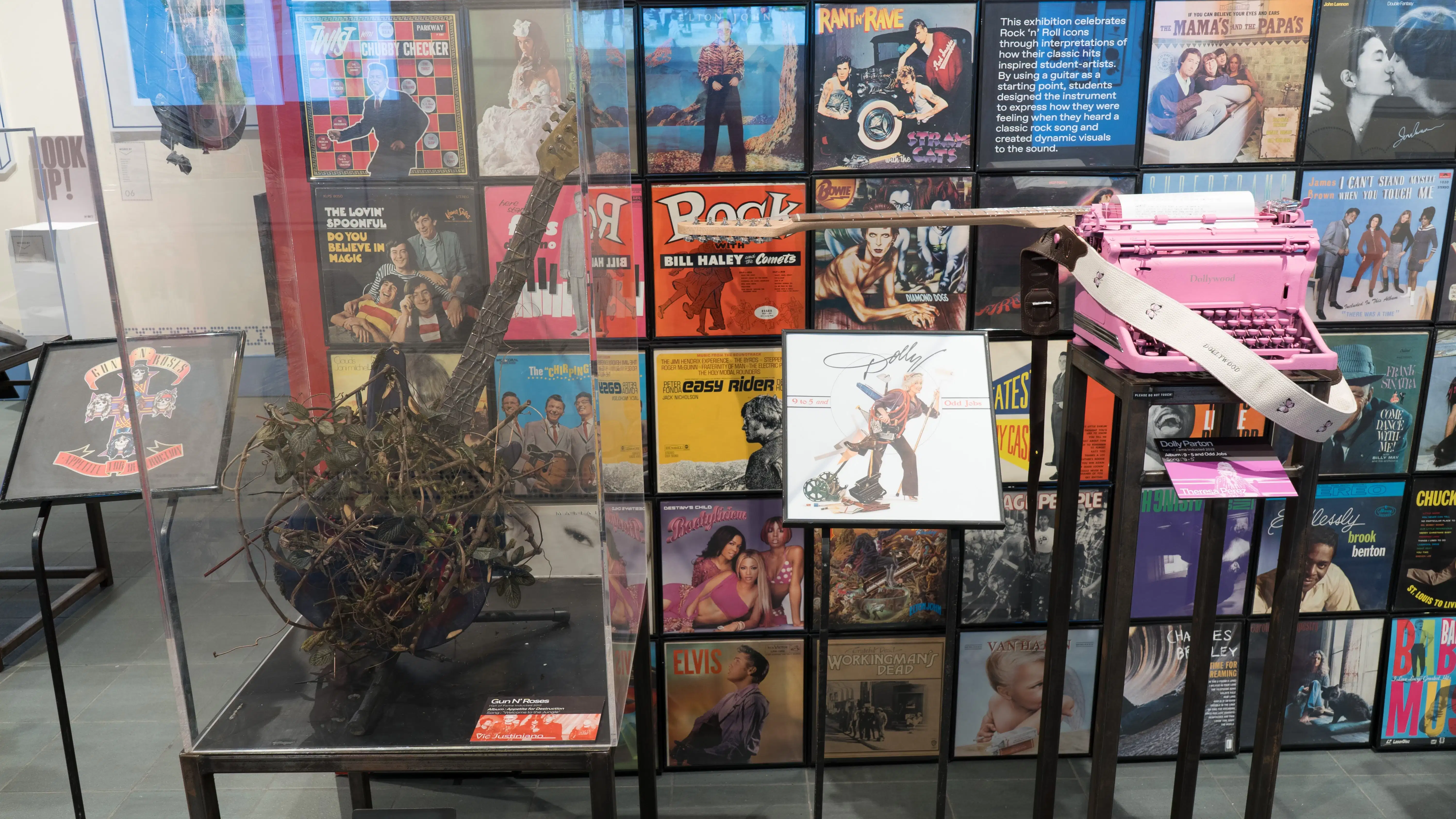 A plaque displaying a Guns N’ Roses album stands next to a guitar, with the body covered in thorny rose bush stems. To the right, a plaque displaying a Dolly Parton album stands next to a guitar made of a pink typewriter.