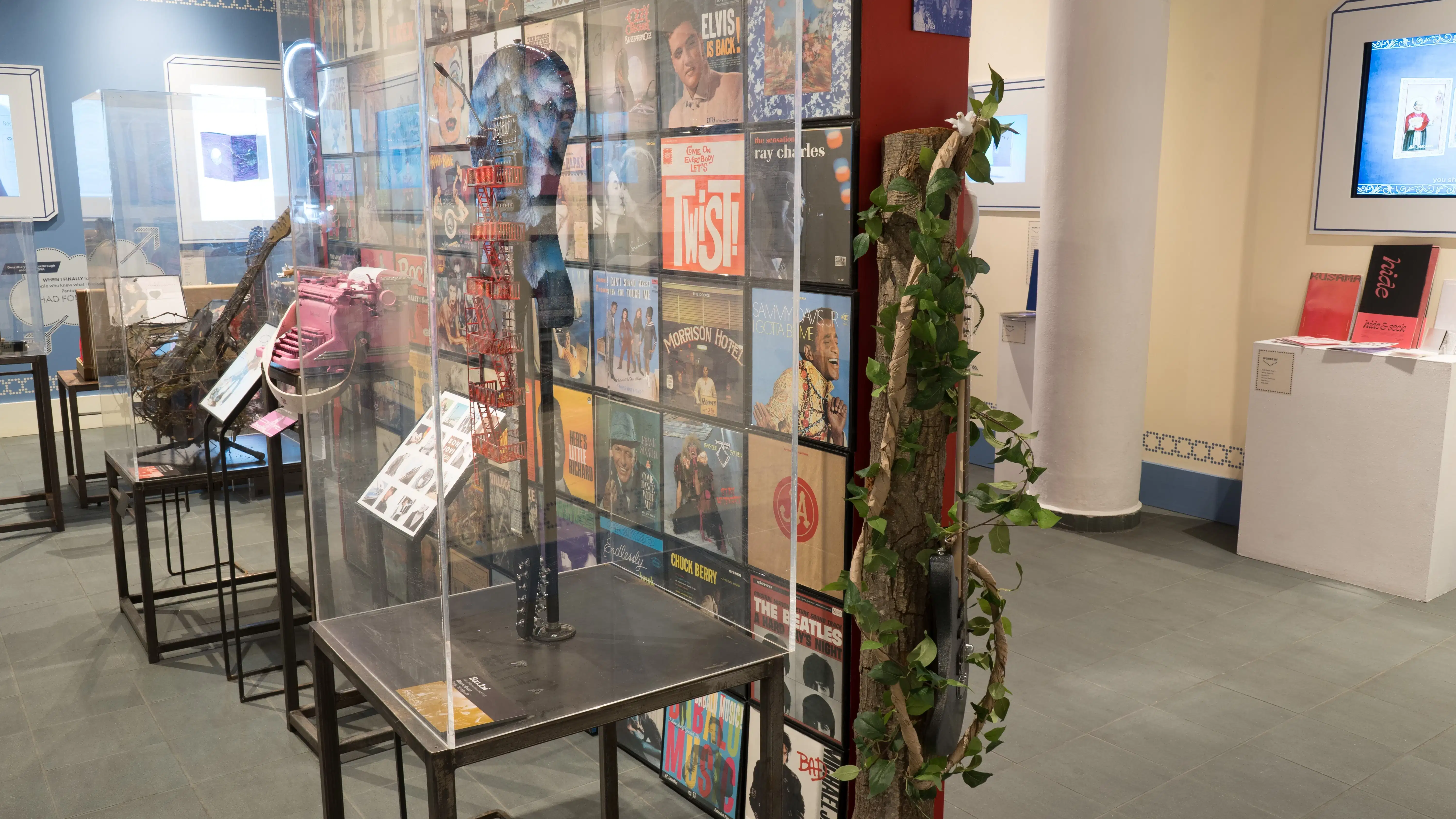 Against a red wall in the center of the room, several unique guitars are shown on pedestals. On the surrounding walls, design and advertising projects play on monitors.