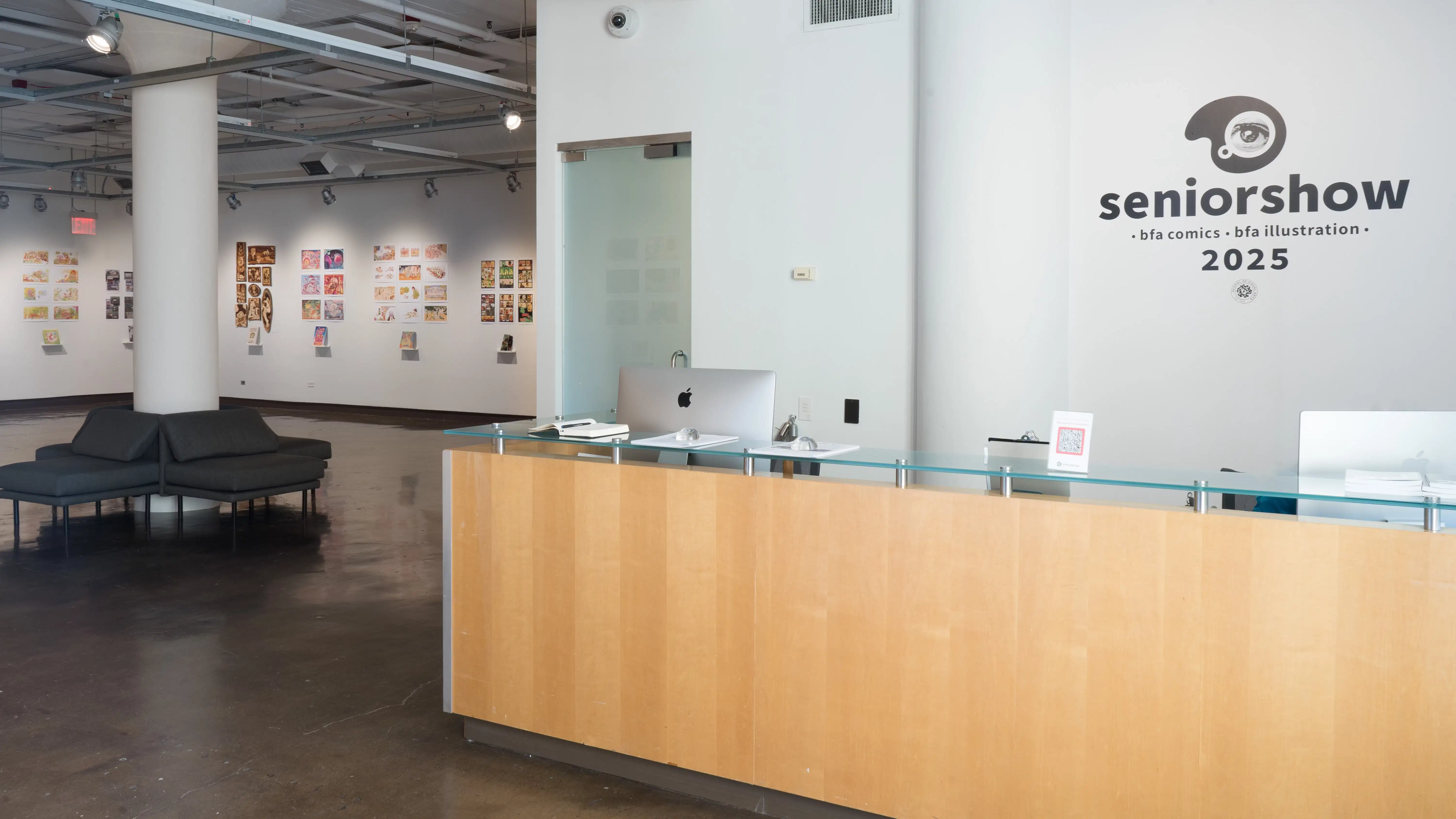 aThe front desk of the gallery sits to the right. On the wall behind the desk, the wall text reads “seniorshow, bfa comics, bfa illustration, 2025”. To the left, several groups of prints are shown, with personalized books displayed under the prints on floating shelves. Four chairs are shown resting alongside a pillar in the main gallery space.