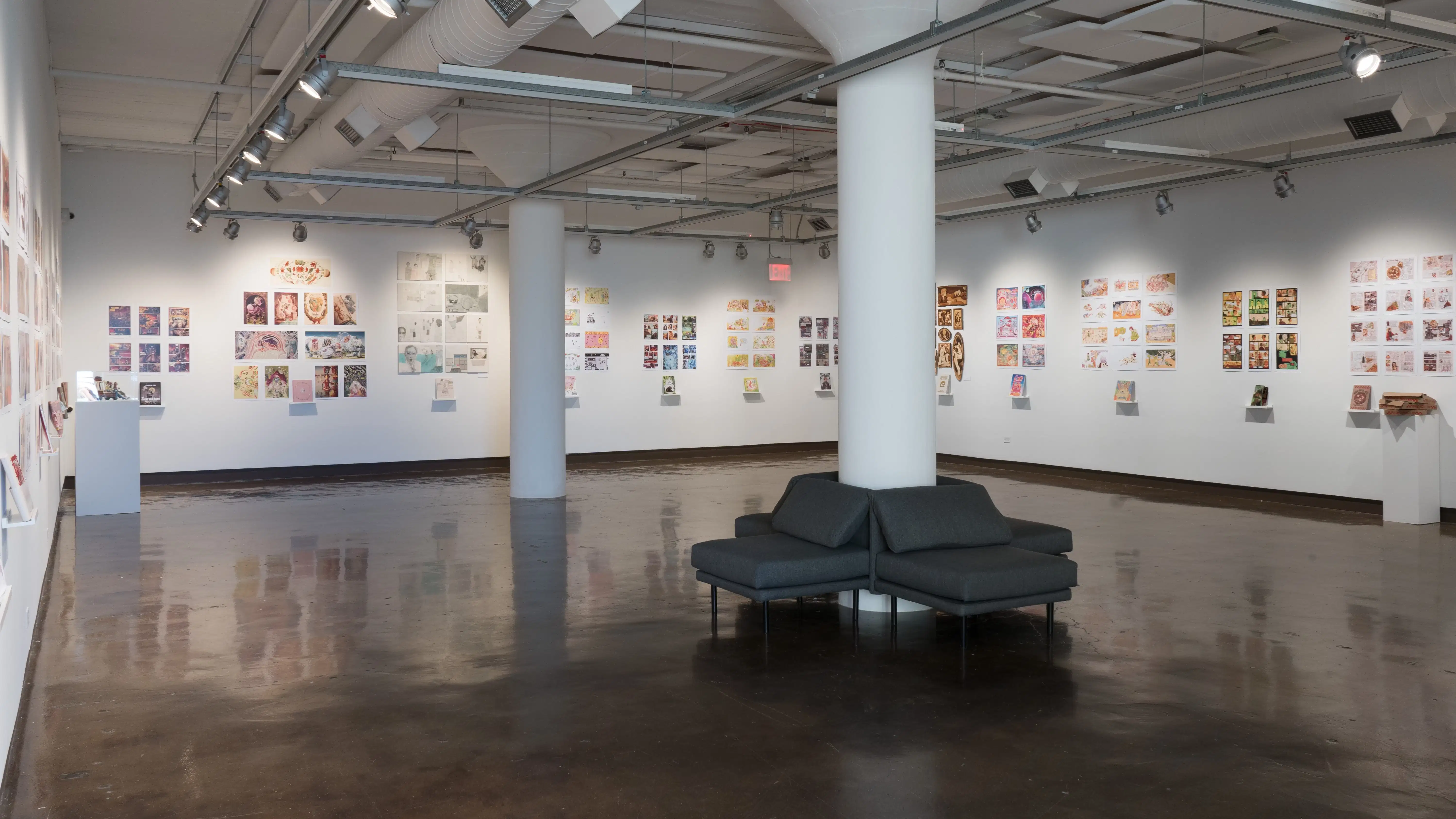 The first gallery space is shown. Three walls are shown, and display several groups of prints all throughout. Each group of prints has a personalized book under the prints on floating shelves. A pedestal sits near the back of the gallery space to the left, with a sculpture of three figures on top the hood of a car. Another pedestal sits near the front on the right, with a sculpture of three pizza boxes, stacked on top of eachother. Two large pillars rest in the middle of the gallery space.